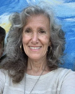 Close-up of a smiling older woman with long gray hair, wearing earrings and a light-colored top, in front of a blue and yellow abstract background.