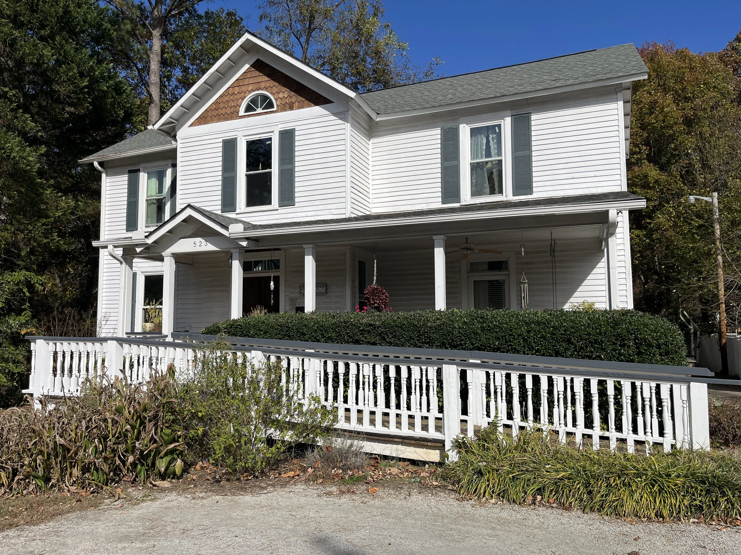 White two-story house with a front porch, gray shutters, and a gable roof, surrounded by trees and a white fence.