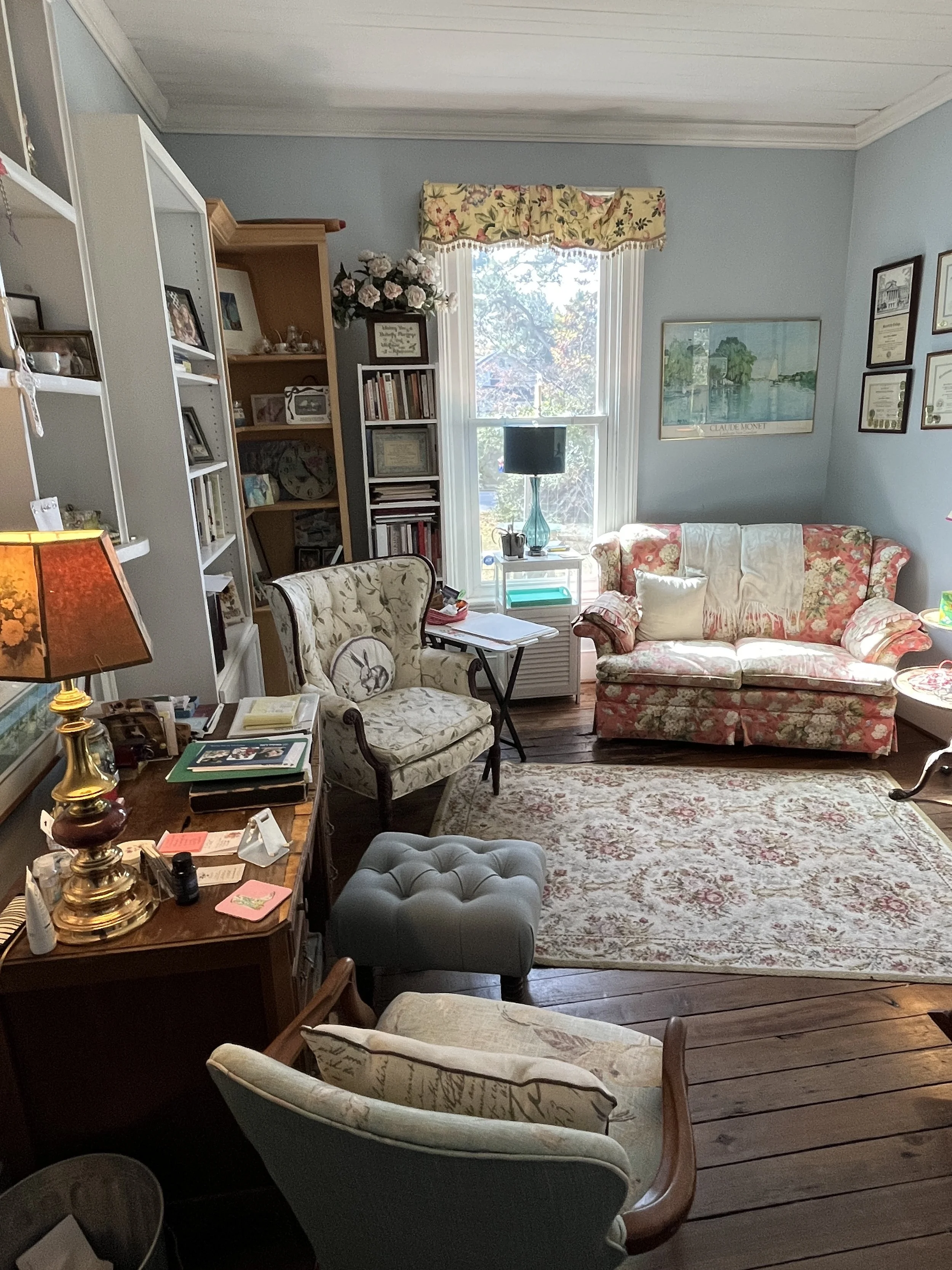 A cozy office space with a floral sofa, armchairs, a rug, bookshelves, and a window with floral valance allowing sunlight to brighten the space.