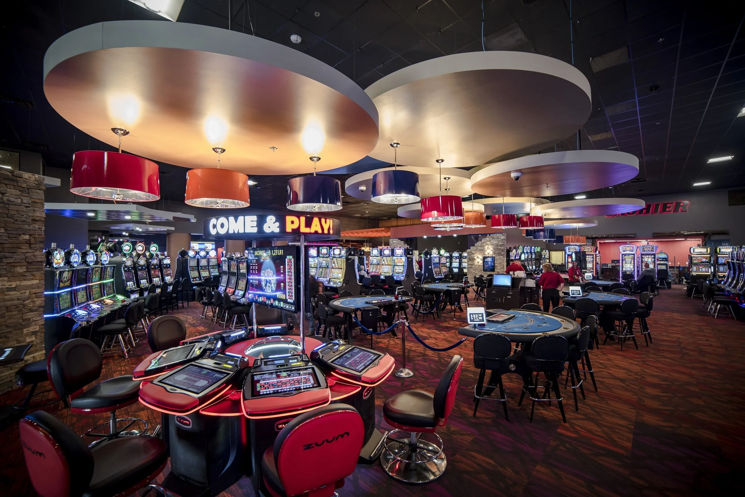 A casino filled with slot machines and electronic gaming tables under a ceiling with large, colorful, circular light fixtures. There is a sign that reads 'COME & PLAY!' with people walking around and gaming activity in the background.