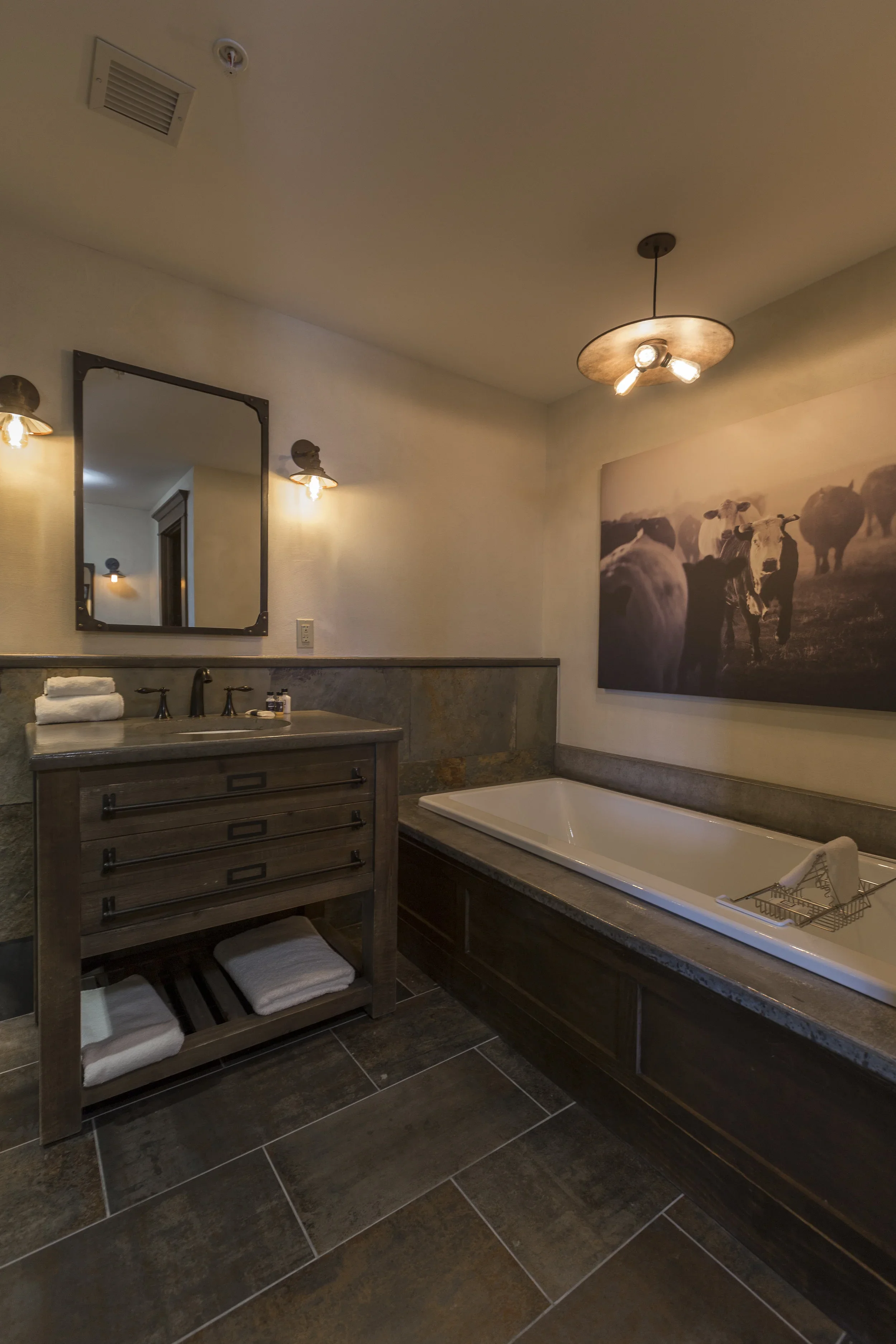 Bathroom with a bathtub, a wooden vanity with a mirror, a large cow-themed wall art, and warm lighting fixtures.
