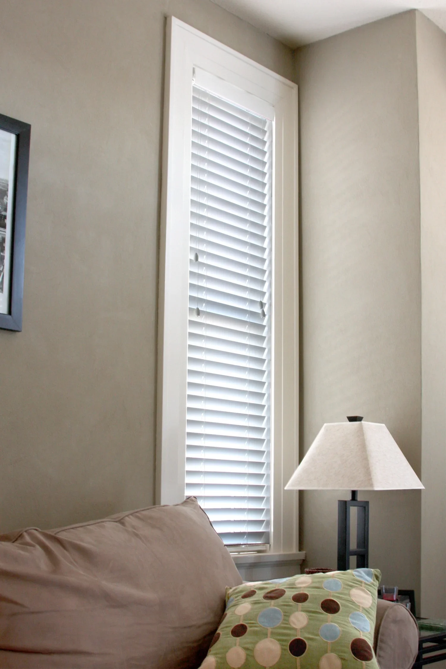 Interior of a living room corner with a beige sofa, a patterned pillow, a table lamp, and a tall window with white blinds.