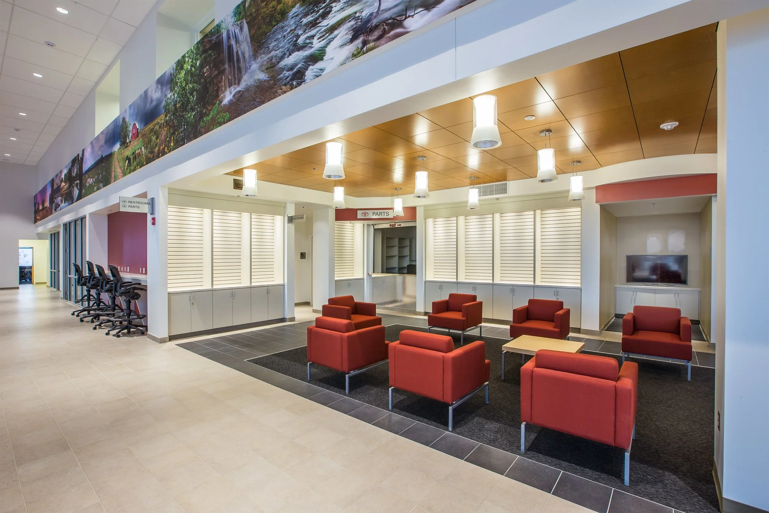 Waiting area with six red armchairs and one beige table, a television, a sign to restroom and parts, and a large mural on the ceiling with nature scenery.