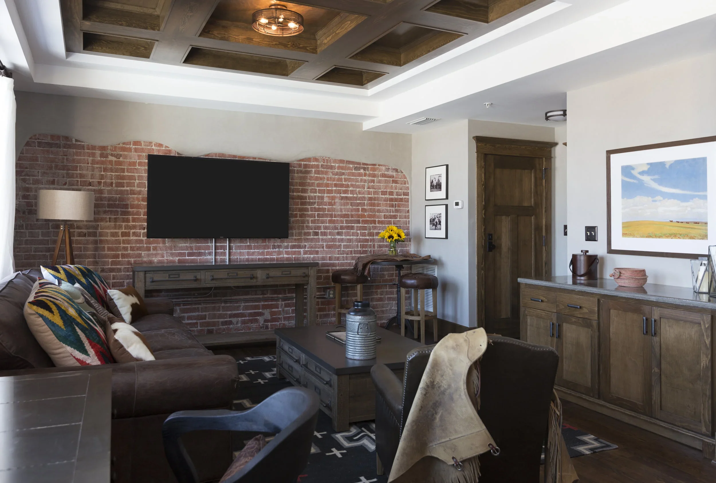 Living room with brick accent wall, mounted flat-screen TV, brown leather sofa with colorful pillows, wooden coffee table, armchair, furniture, framed pictures, window with white curtains, wooden door, and art on the wall.