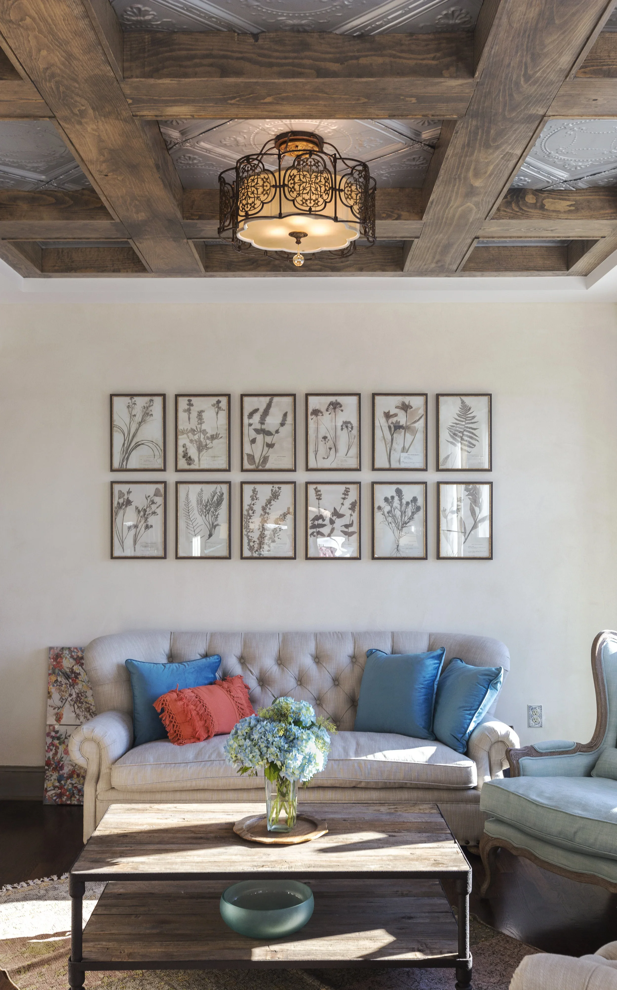 Living room with a beige tufted sofa with blue and red pillows, a wooden coffee table with a vase of blue hydrangeas, framed botanical artwork on the wall, a chandelier, and a wooden ceiling with decorative white tiles.