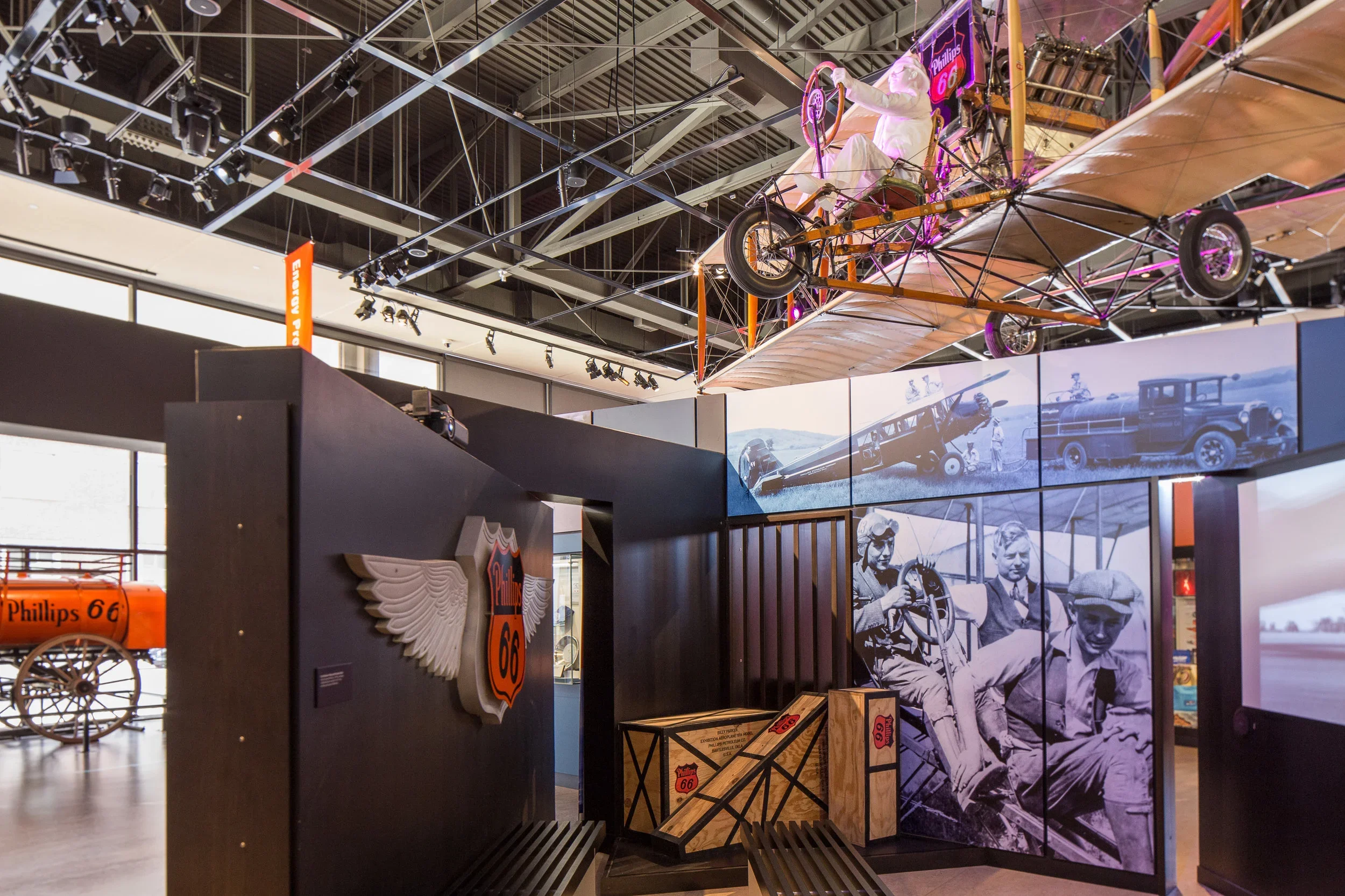 Museum exhibit featuring a model of a vintage biplane with a human size figure sitting on it, vintage black and white photos of early aviation and automobiles on the walls, Route 66 emblem with wings on the wall, and a wooden crate with Route 66 bran