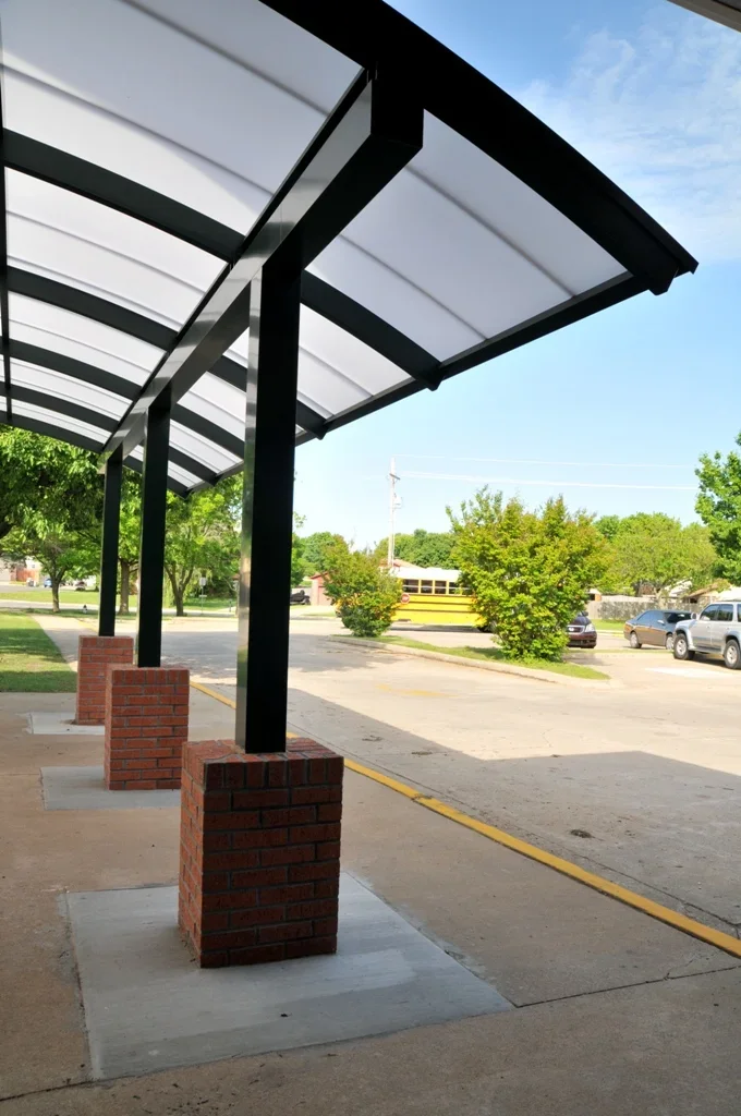 Empty sidewalk with a covered walkway supported by black metal posts and red brick bases, next to a parking lot with a school bus, cars, trees, and a blue sky.