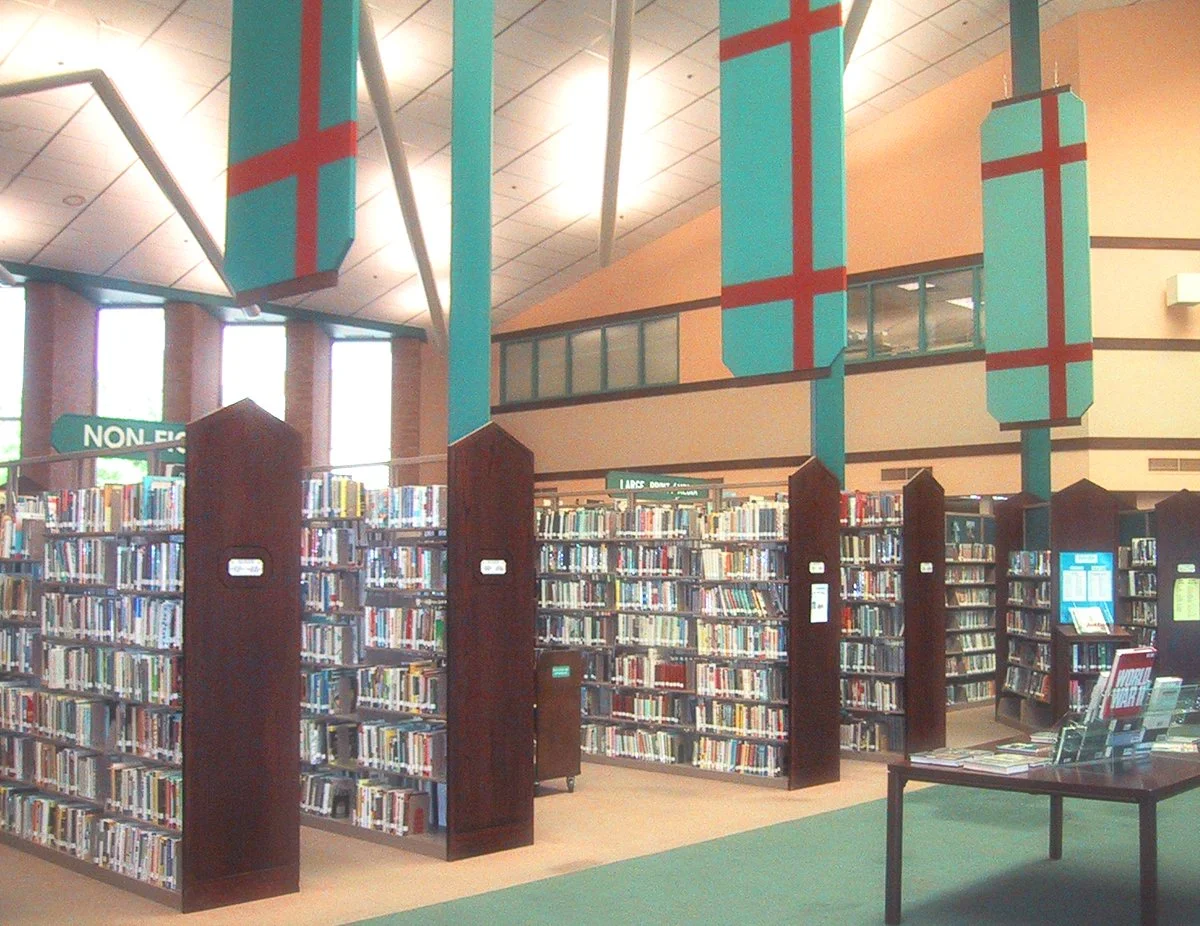 View of a public library with multiple bookshelves filled with books. The ceiling has large hanging decorations with a blue background and a red cross pattern. There are signs indicating sections and a table with informational pamphlets.