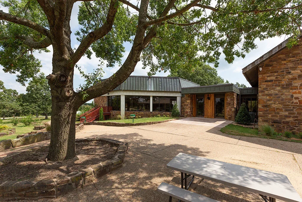 A large tree with a thick trunk and green foliage in front of a building with large windows, brick walls, and a metal roof. A concrete pathway leads to the building entrance, with a picnic table and a landscaped yard surrounding it.