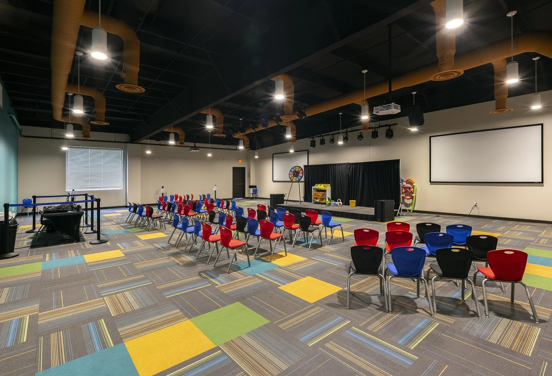 An empty conference or presentation room with colorful chairs arranged in rows facing a stage with black curtains, large screen, and various equipment, including a spinning wheel and toys, with multicolored patterned carpet and ceiling lights.