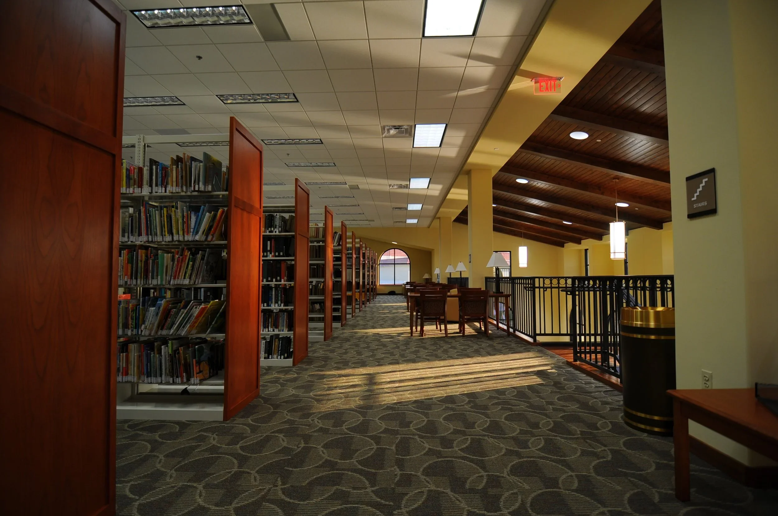 Interior of a library reading area with bookshelves, seating, and large windows, illuminated by natural and artificial light, featuring a carpeted floor and wooden ceiling.