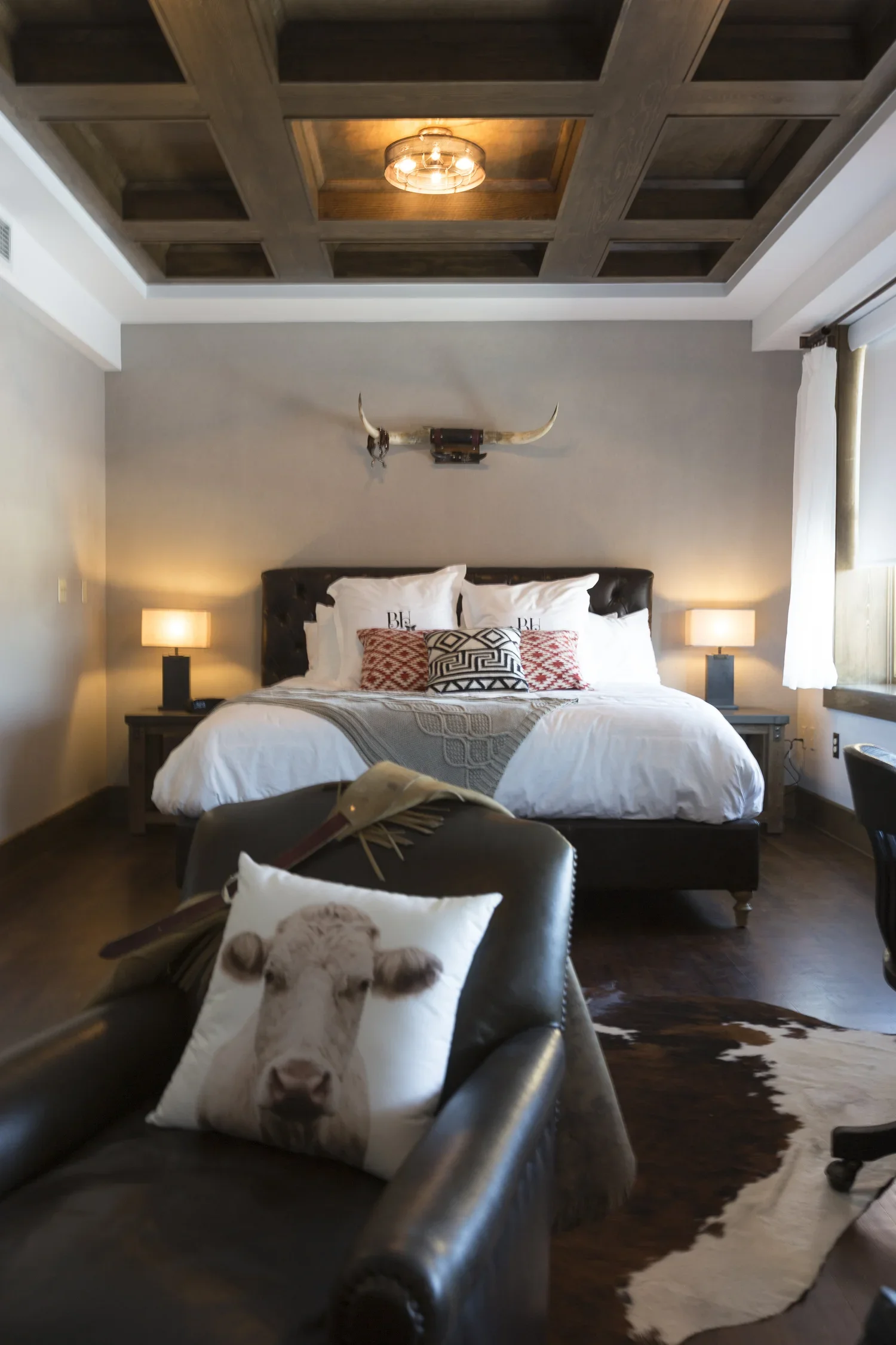 A bedroom with a large bed, decorative pillows, bedside lamps, a mounted bull horn, and a cowhide rug on the floor. There is a pillow with a cow face on the armchair in the foreground, and the ceiling has a coffered wood design with a modern light fi