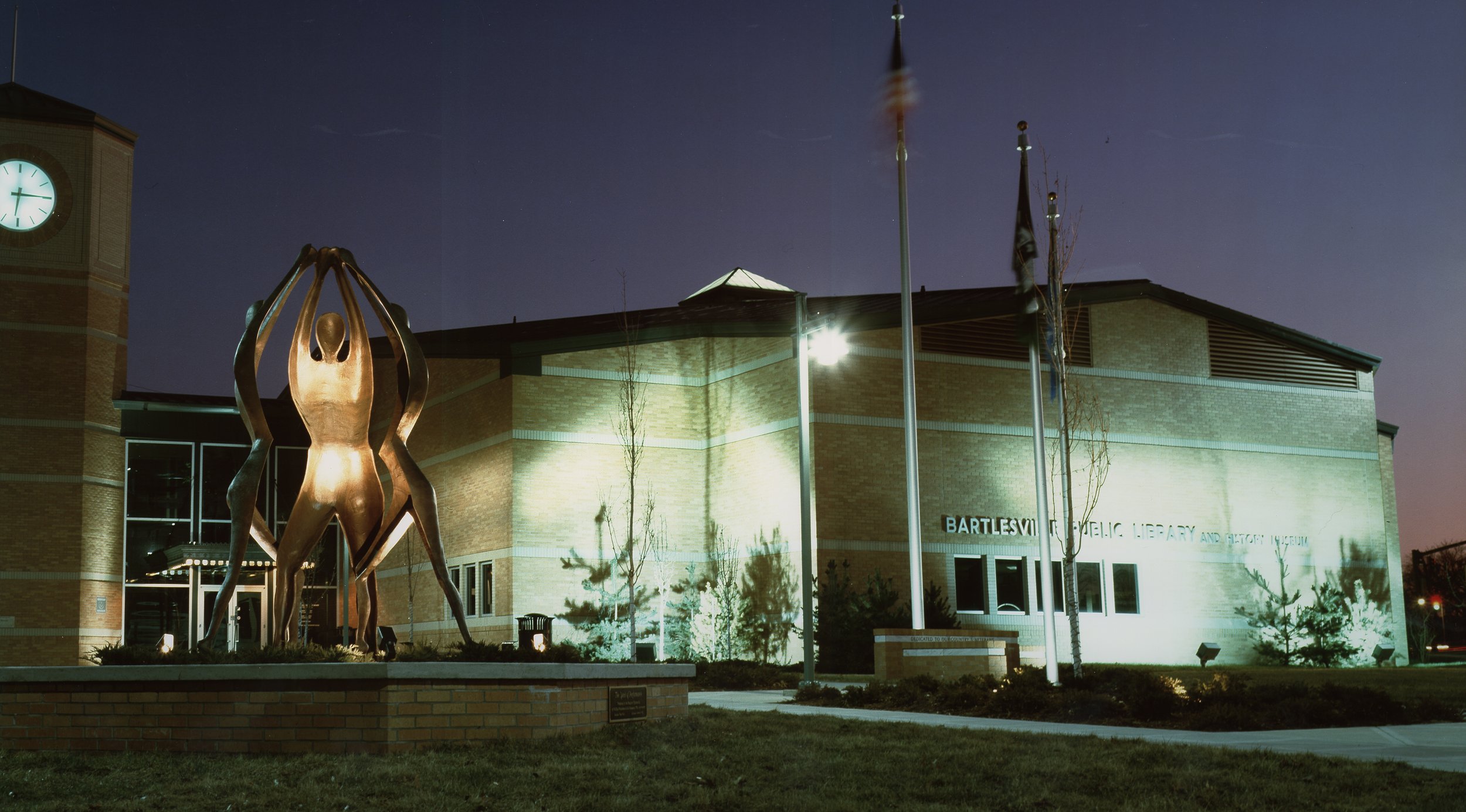 Nighttime exterior view of a library building with a sculpture of human figures in front, illuminated by exterior lights, with flags on tall poles nearby.