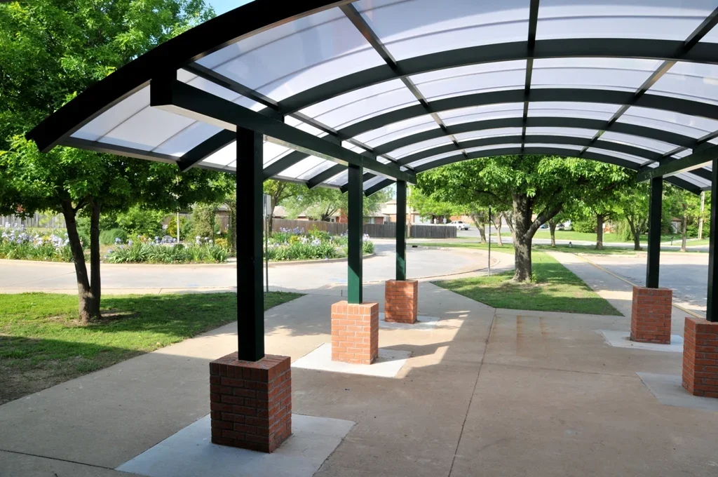 Covered outdoor pavilion with metal roof supported by brick columns, situated on a sidewalk with trees and greenery in the background.