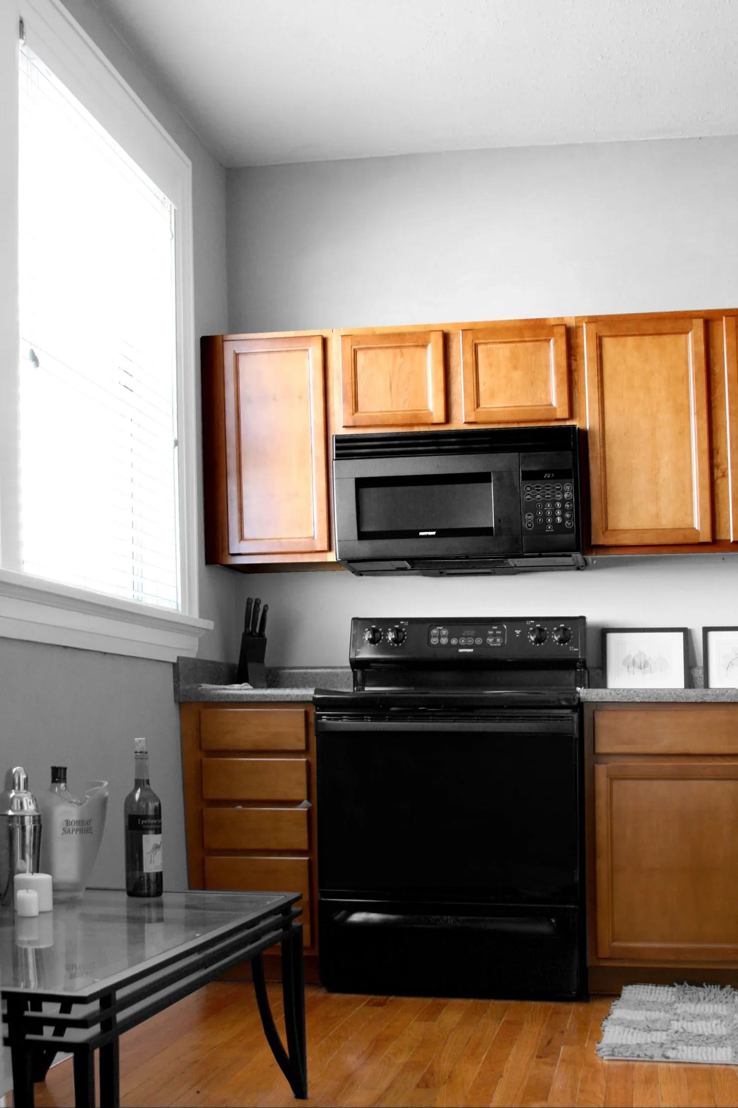 A kitchen with wooden cabinets, a black stove and microwave, a window on the left letting in natural light, a countertop with a knife block, framed pictures, and bottles on the counter, and a small table with drinks and candles in the foreground.