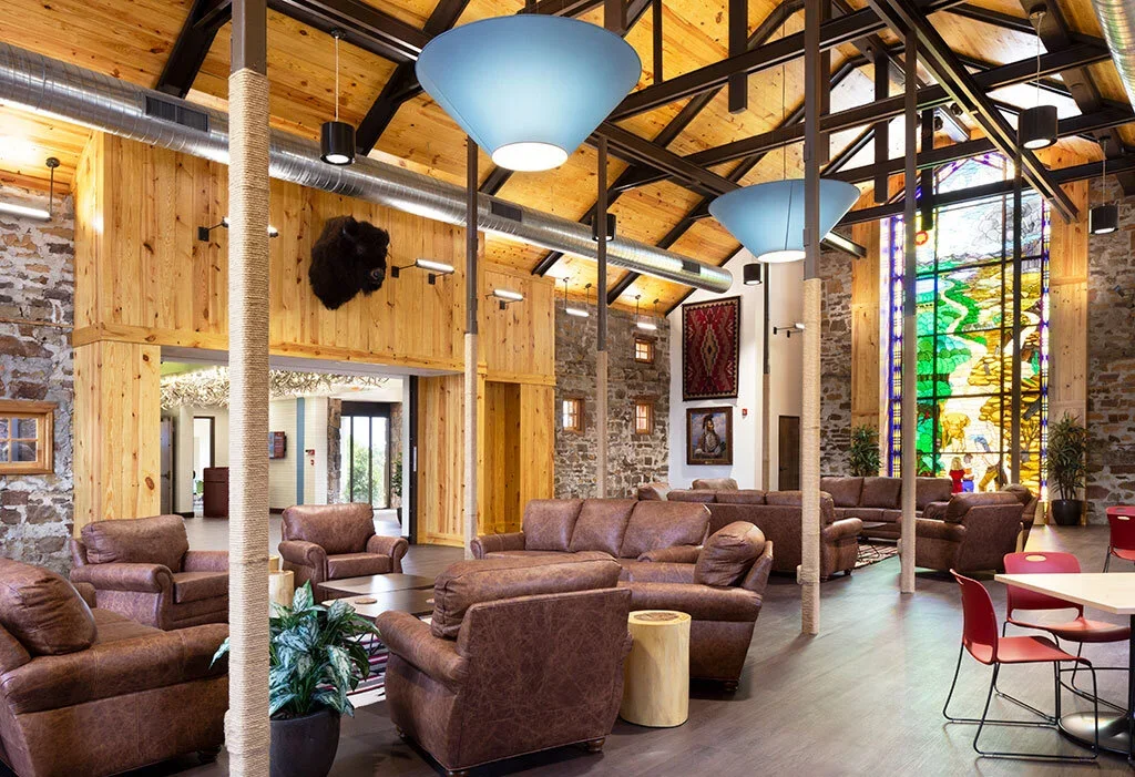 A spacious lounge area with leather sofas, colorful hanging lamps, a large stained glass window, and wooden accents, inside a modern rustic building.