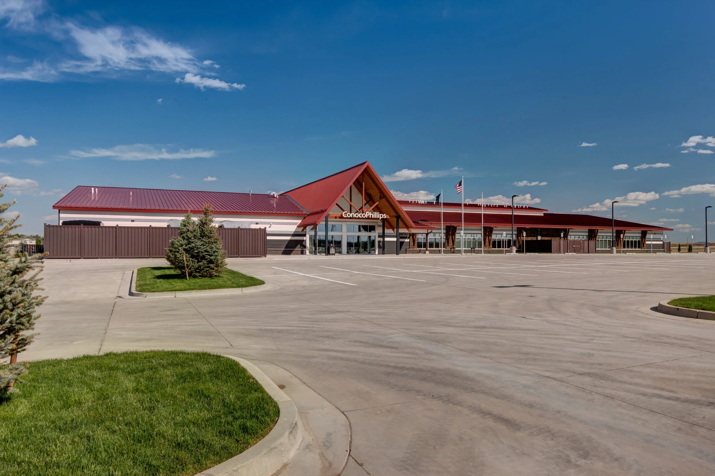 Empty parking lot in front of ConocoPhillips building with flags, small trees, and manicured grass under a blue sky with clouds.