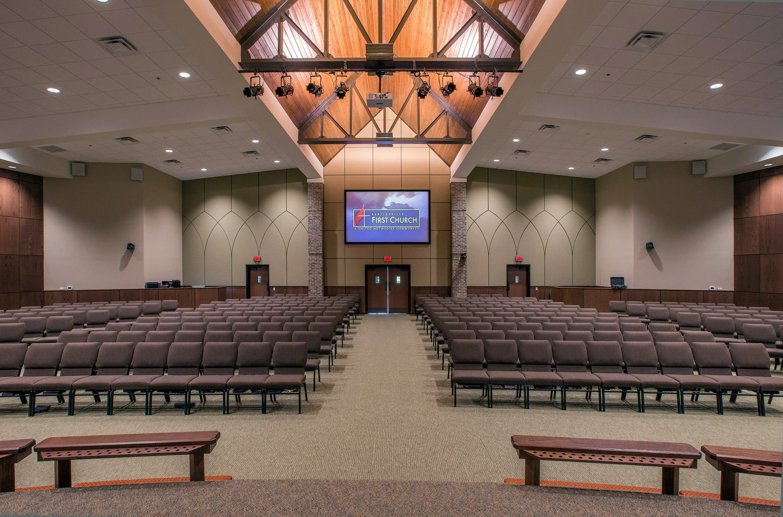 An empty church auditorium with rows of brown chairs facing toward a stage with a large screen, wooden ceiling, and brick pillars, labeled 'First Church'