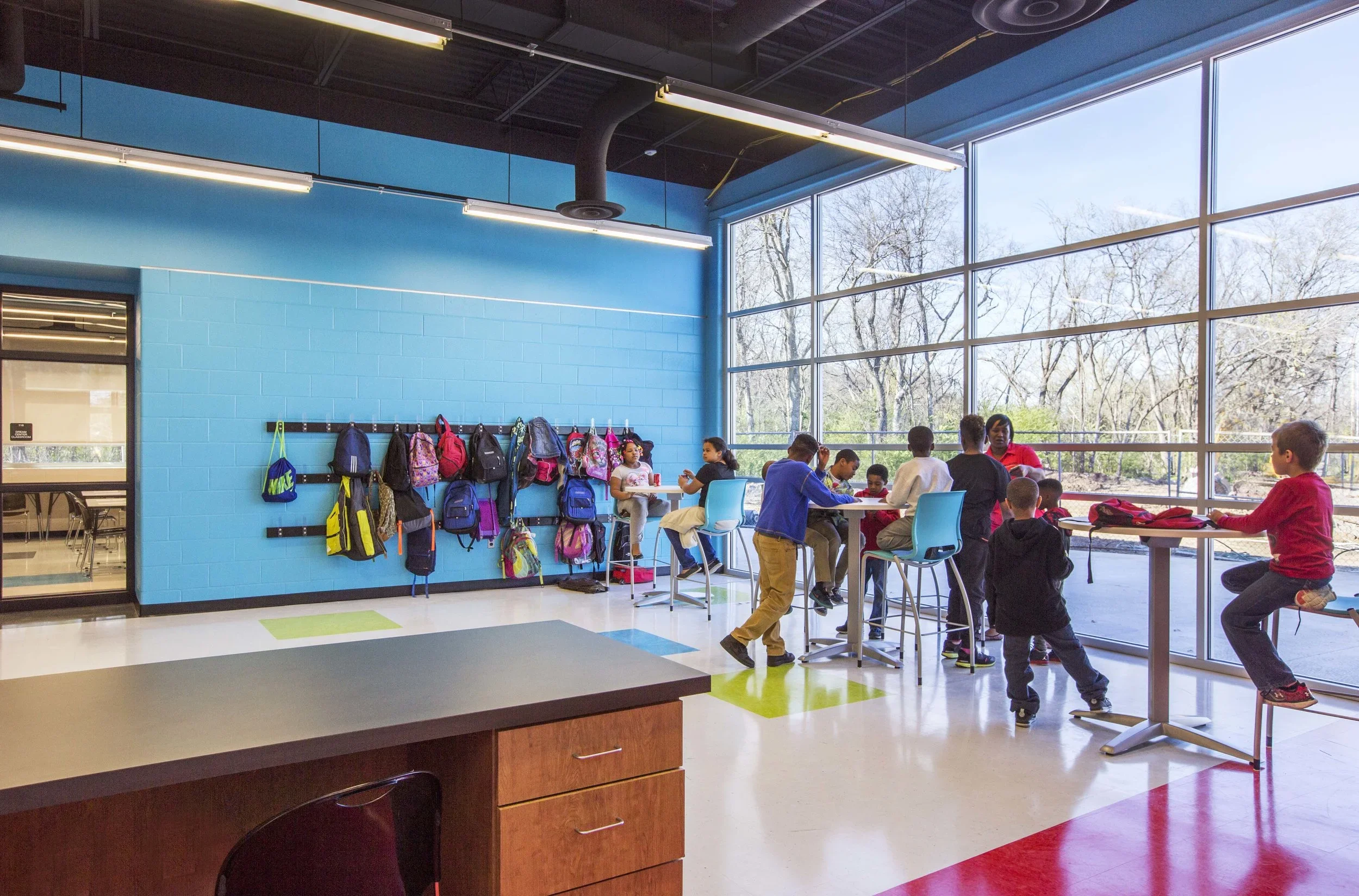 Children in a classroom with large windows, some sitting at tables, others on stools, backpacks hanging on hooks on a blue wall, bright interior with outdoor view.