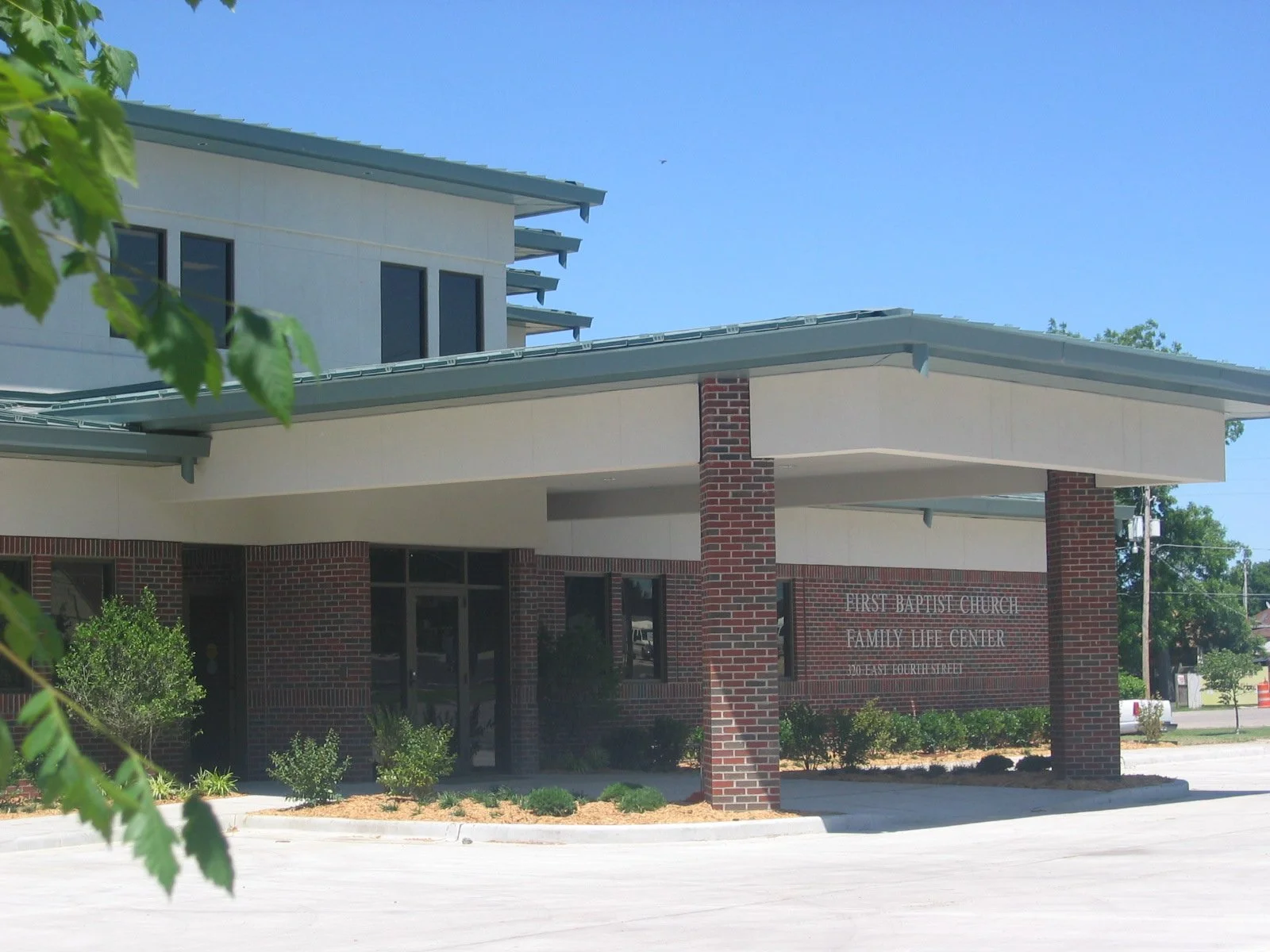 Exterior view of First Baptist Church Family Life Center, a modern building with brick and light-colored walls, surrounded by landscaped bushes, under a clear blue sky.