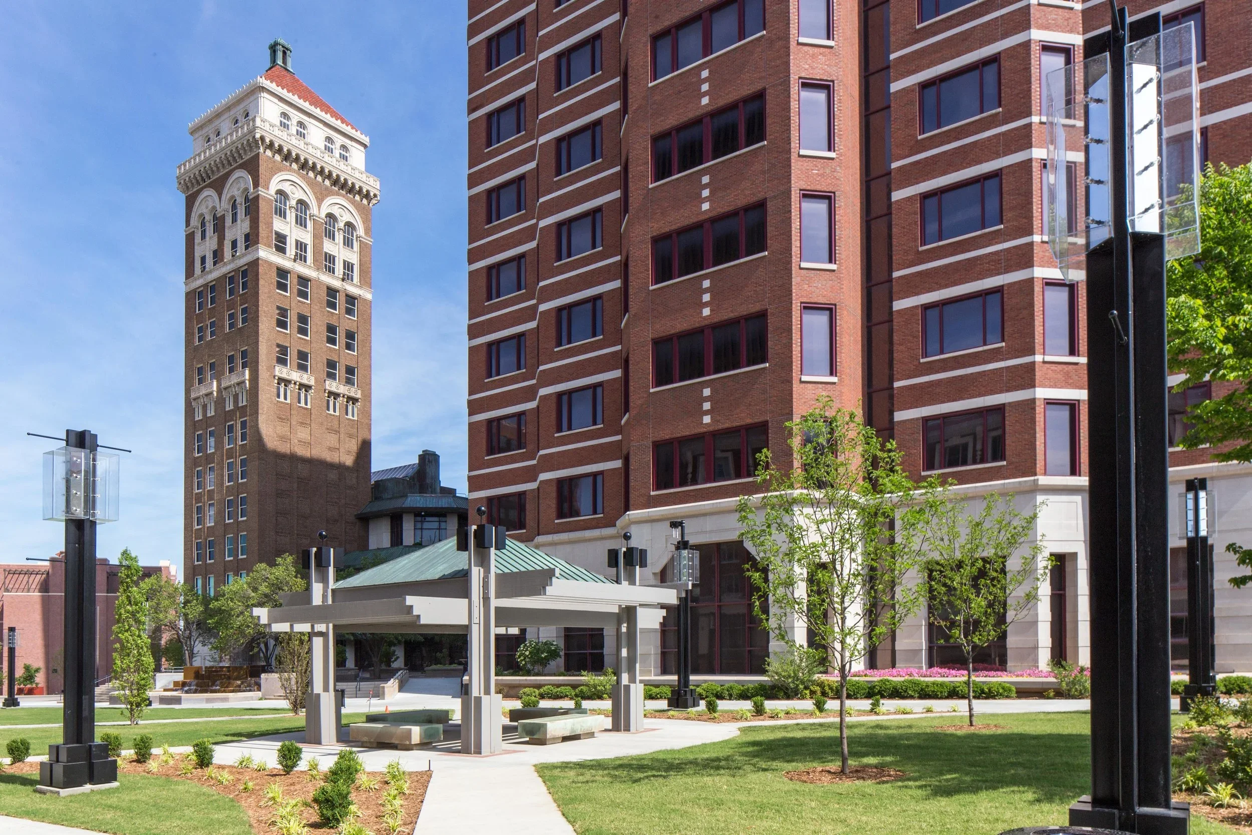 A city park with a green lawn, young trees, and a white pavilion in front of tall brick buildings, including a historic clock tower, on a clear day.