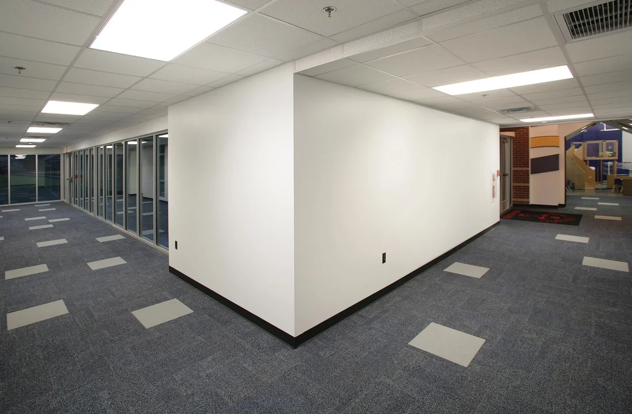 Empty indoor office corridor with white walls, gray carpeted floor with white square tiles, glass-walled rooms on the left, and a children's play area in the background on the right.