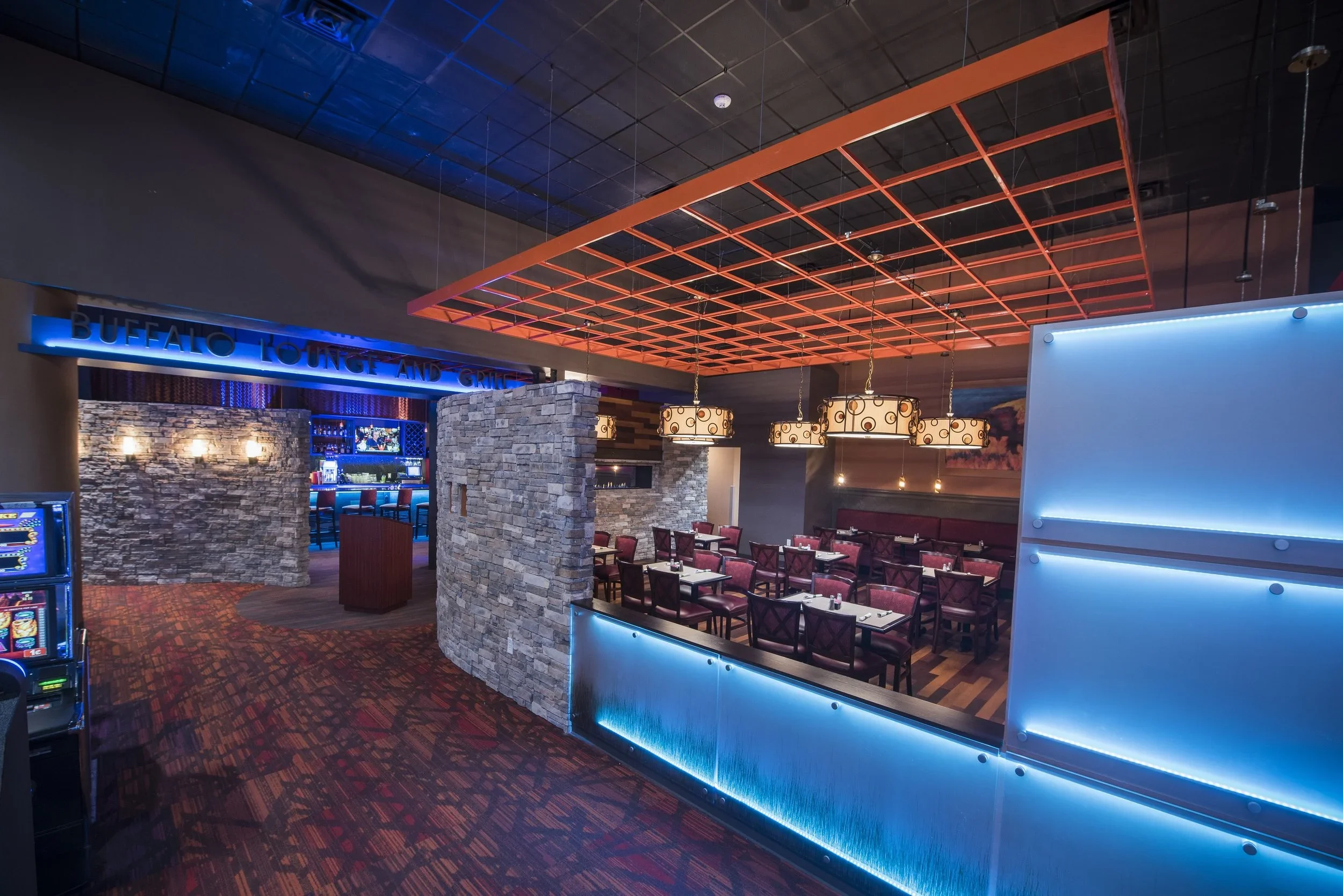 Interior of a restaurant with red chairs, stone walls, hanging pendant lights, a blue neon sign reading 'Buffalo Lounge and Grill', and a ceiling with orange grid design.
