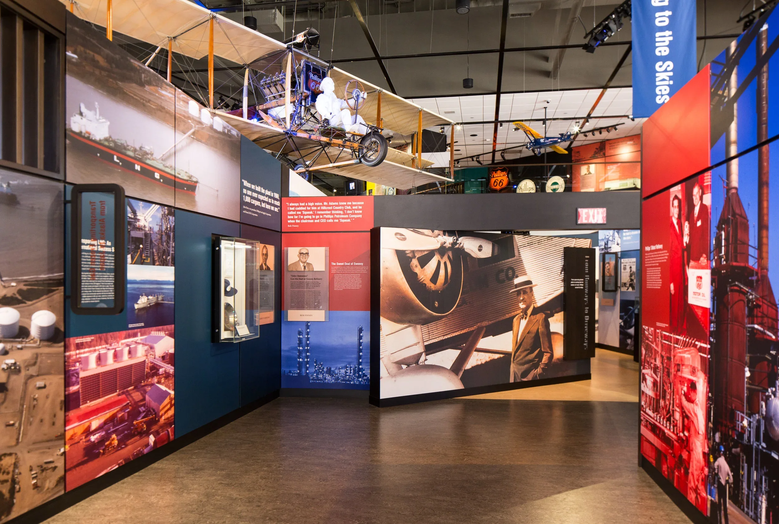 Interior of a museum exhibit with vintage aviation and industrial images, a hanging wooden airplane and a vintage manikin, and various informational panels.