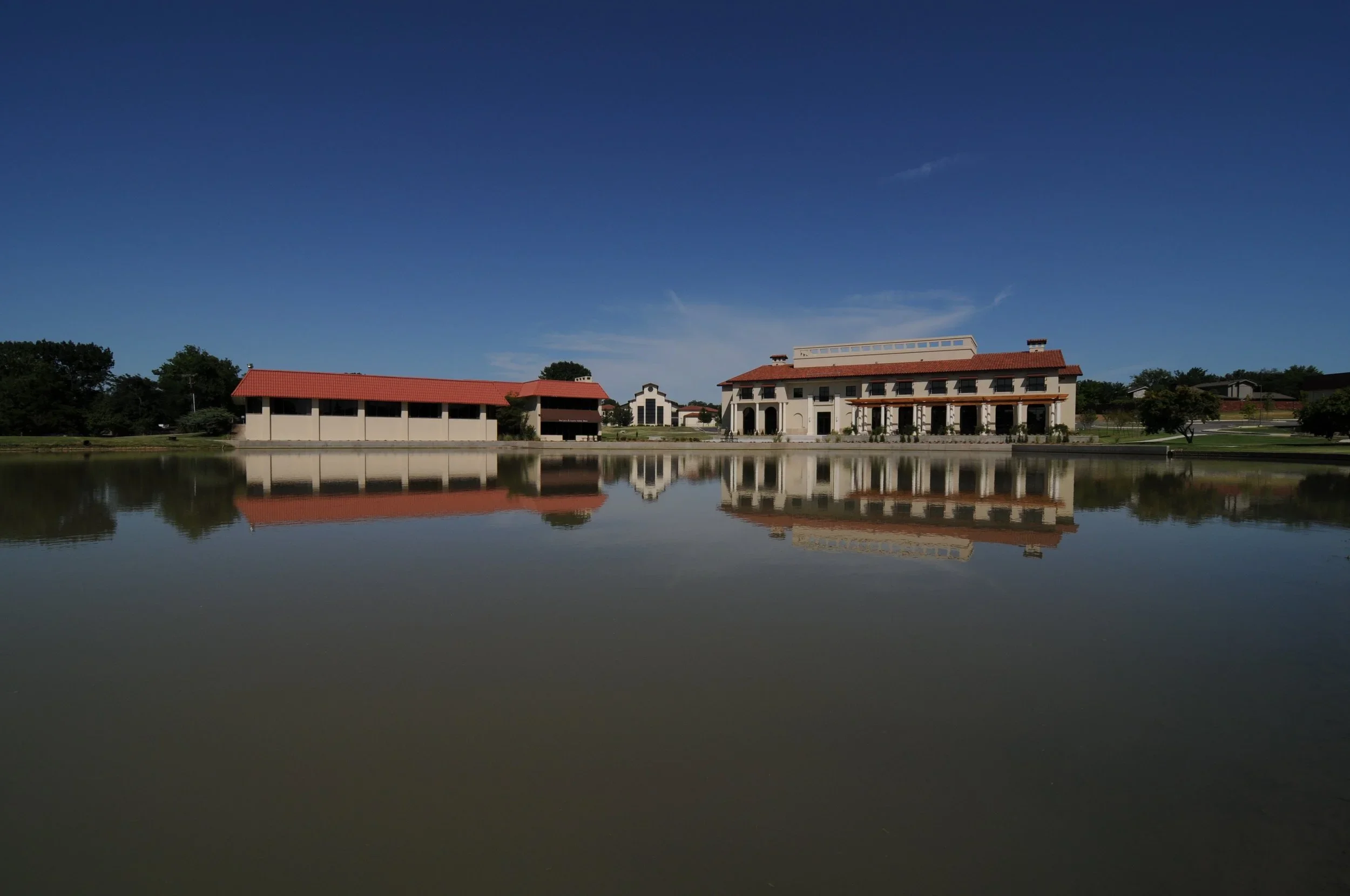 A large building with a red roof and white walls is reflected in a body of water in front of it. The sky is clear and blue.
