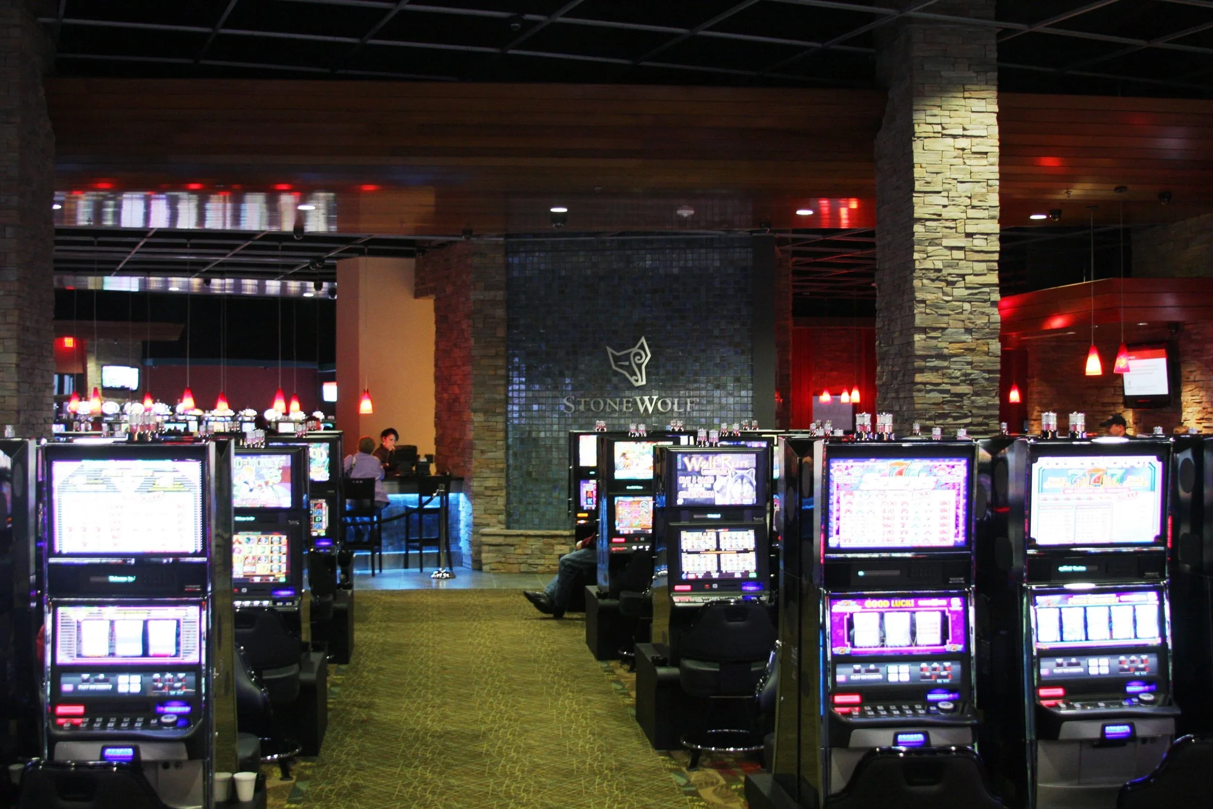 Casinos with slot machines and blackjack tables, illuminated with red hanging lights and dark stone walls, in the Stone Wolf casino.