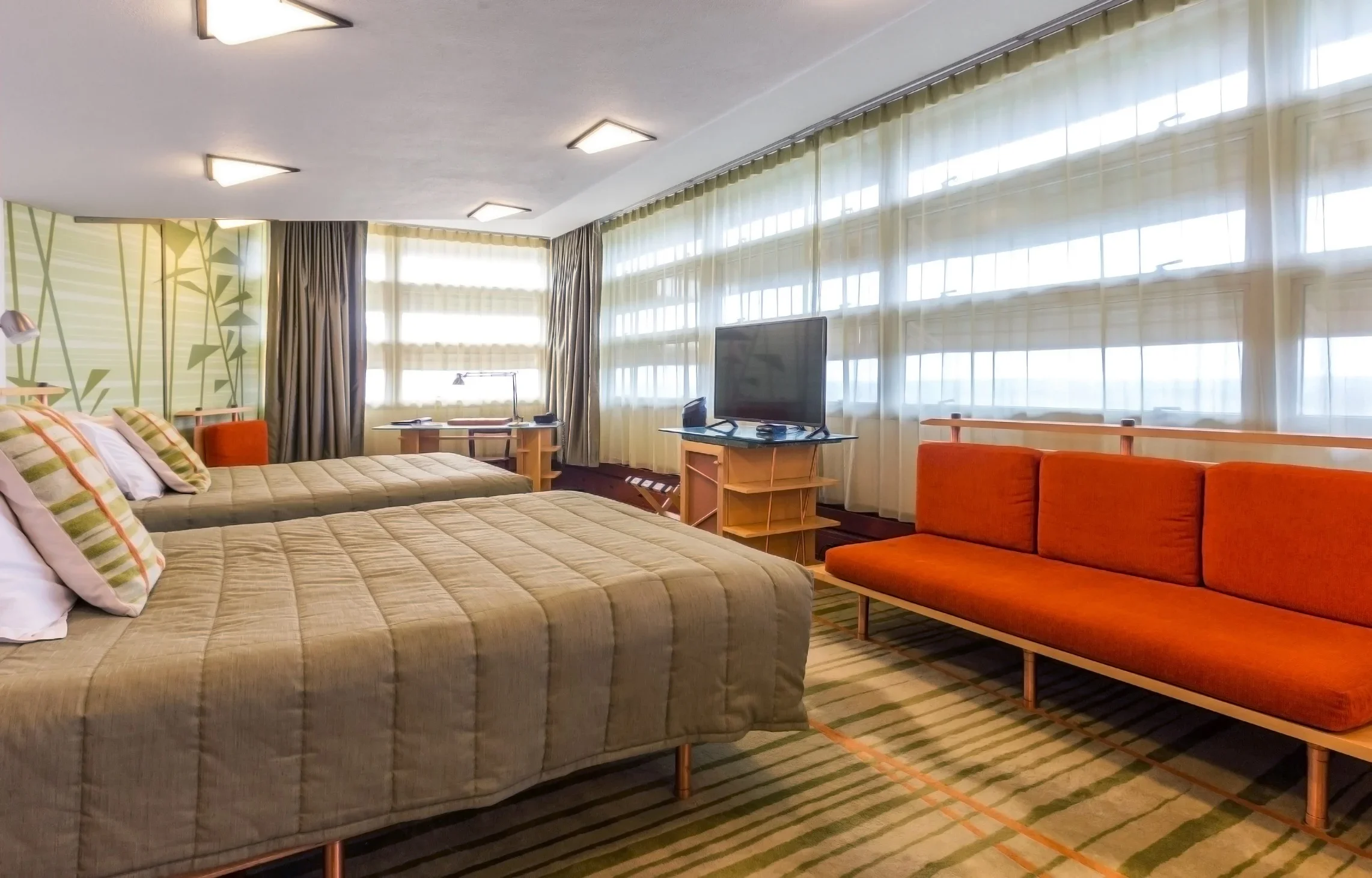 A hotel room with two beds, a bright orange sofa, a flat-screen TV on a wooden stand, large windows with sheer curtains, a desk with a lamp and chairs, and a patterned carpet.