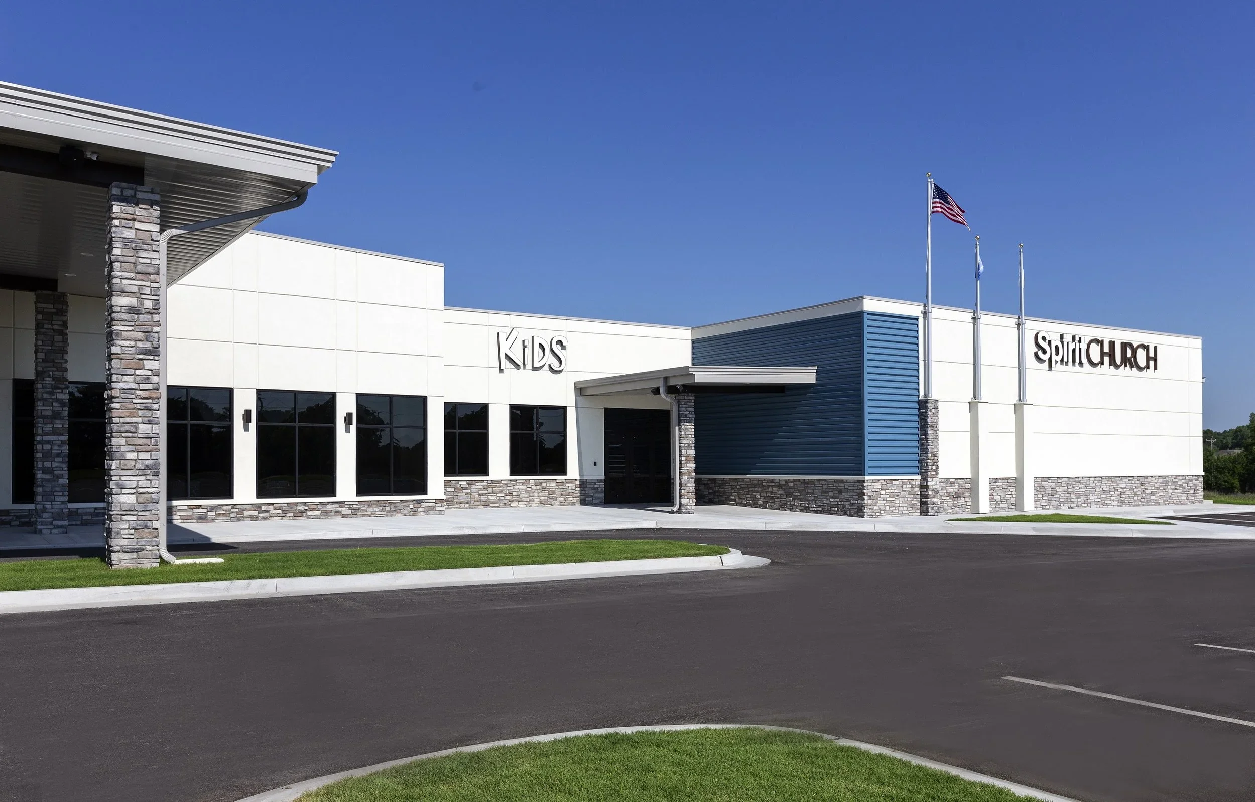 Modern church building with a sign reading "Kids" and "Spirit Church" on the exterior. The building has white and blue exterior walls with stone accents, large windows, and flags flying on poles outside. The area has a parking lot and green grass.