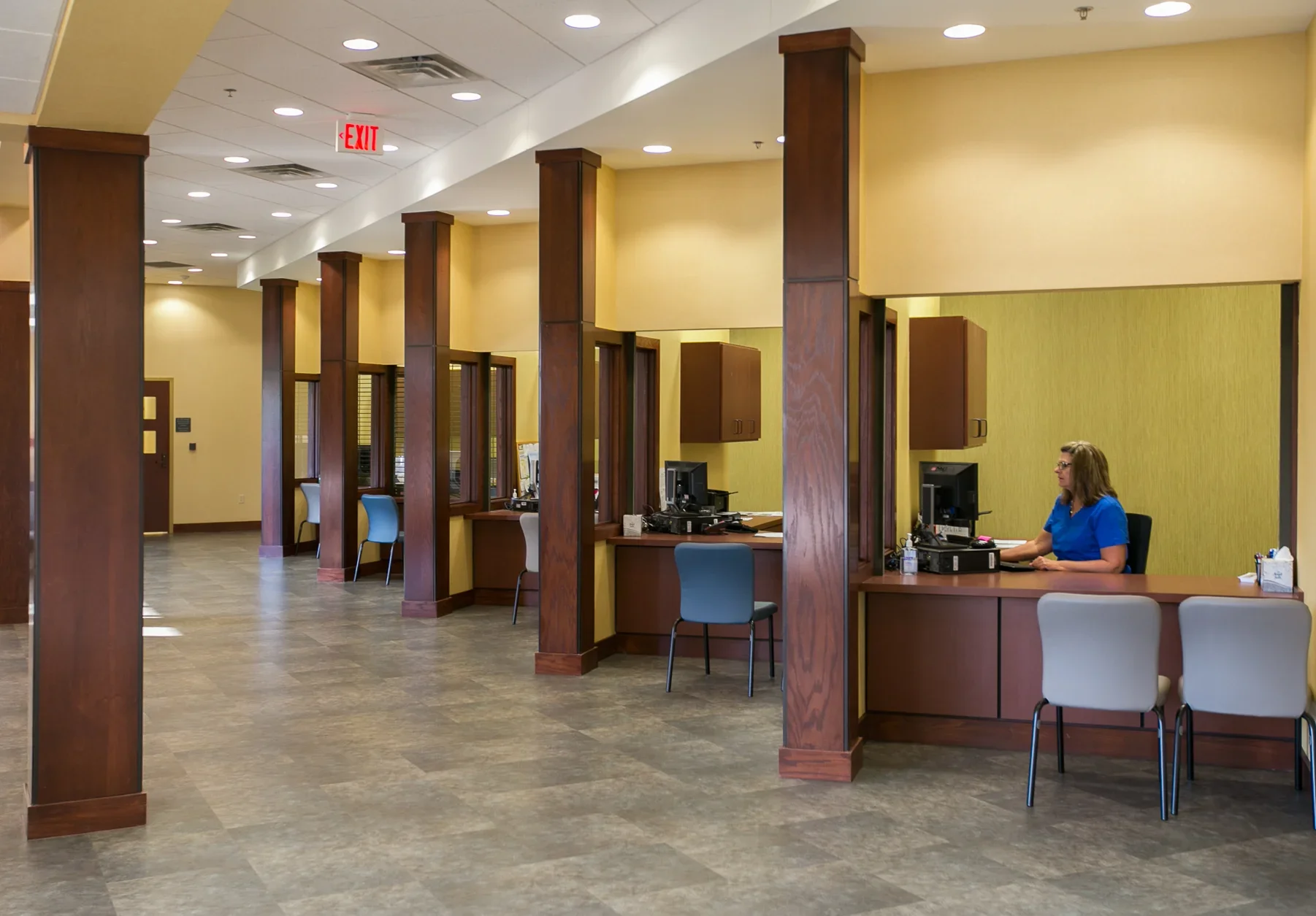 A bank or medical office reception area with four customer service desks, each with a computer and chair, separated by wooden partitions, a woman in medical scrubs working at one desk, yellow walls, wood accents, and an exit sign overhead.