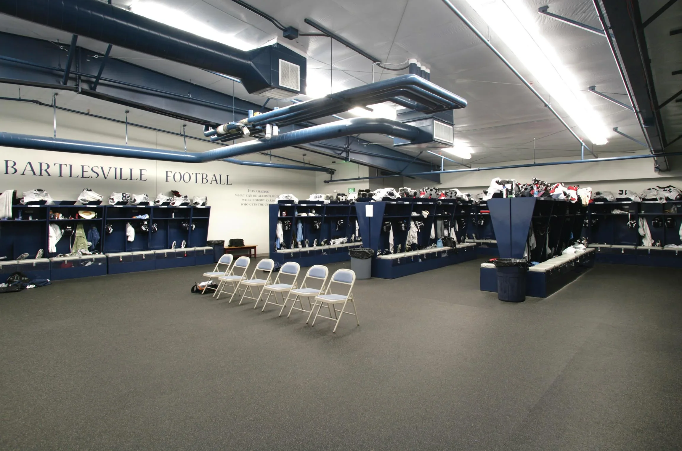 A football locker room with blue lockers, football gear, and chairs in the center.