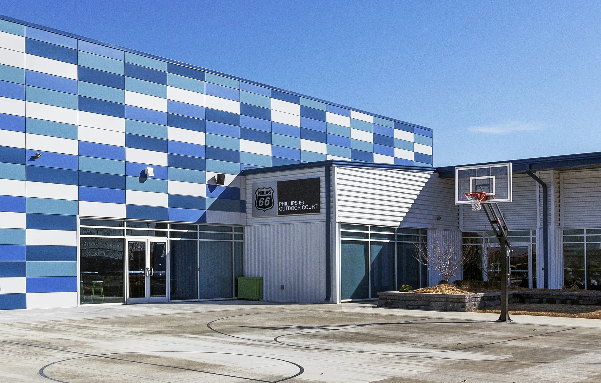 An outdoor basketball court with a hoop and backboard, adjacent to a modern building with a blue and white patterned exterior and a sign that reads 'Phillips 66 Outdoor Court.'