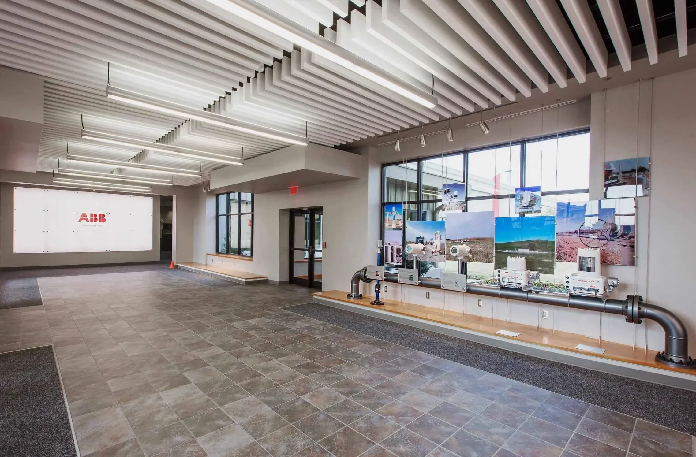 Modern office lobby with gray tiled floor, large windows, and a display area with industrial pipes and photos. An illuminated white wall with the ABB logo is visible in the background.