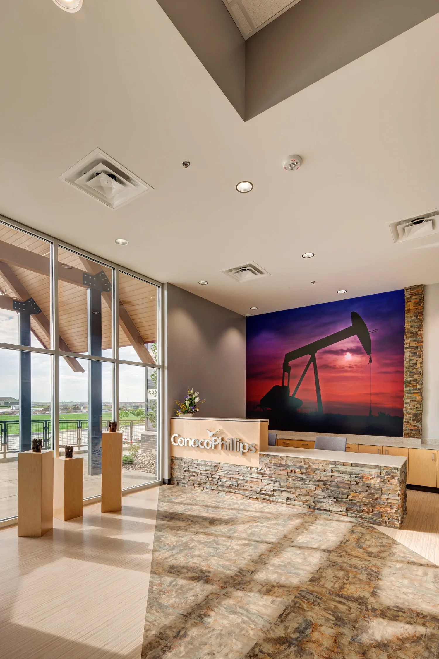 Office reception area with a stone front desk displaying the ConocoPhillips logo, large window with a view of green fields, a mural of an oil pumpjack at sunset on the wall, and a small arrangement of flowers on the desk.