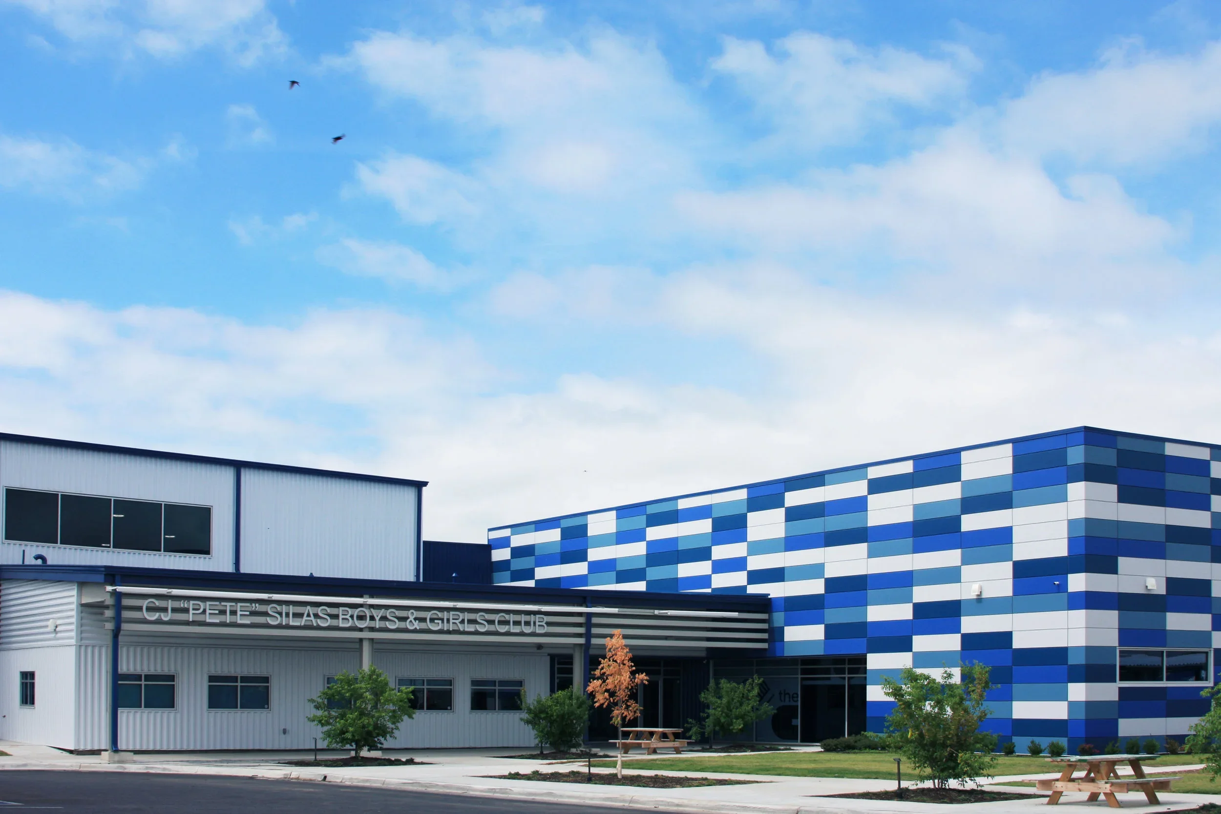 Exterior of the CJ 'Pete' Silas Boys & Girls Club building with a blue and white checkered facade, trees, a picnic table, and a blue sky with clouds and birds