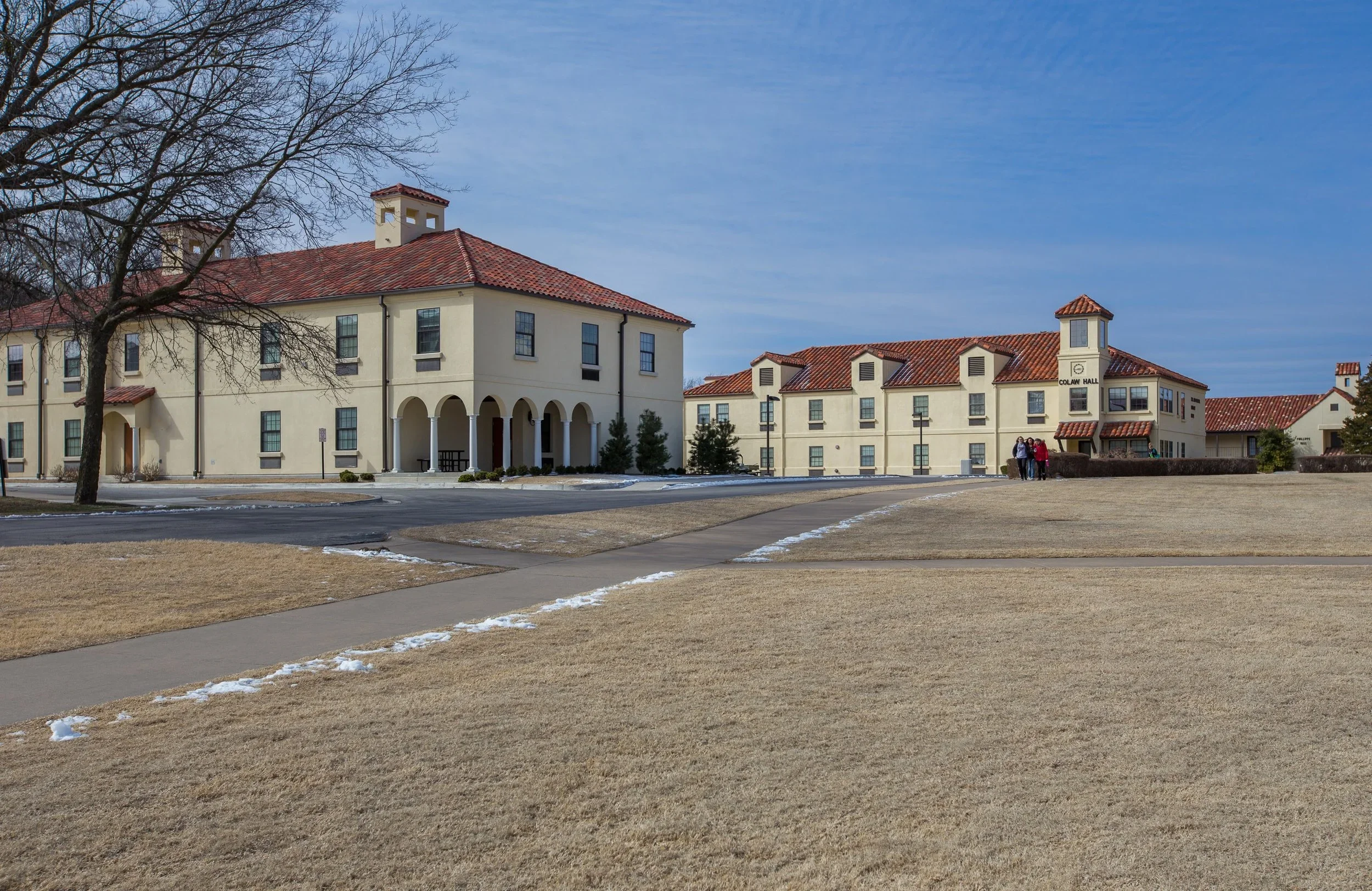 A college campus with beige buildings, red-tiled roofs, leafless trees, sidewalk paths, and a group of people walking near a building labeled 'Colaw Hall' on a partly cloudy day.