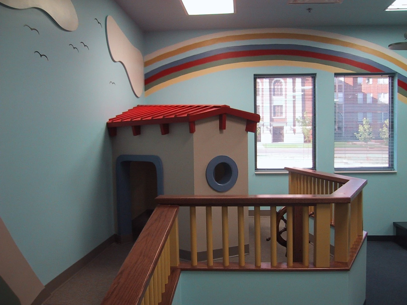 Colorful indoor children’s play area with a small playhouse, rainbow mural, and windows showing buildings outside.