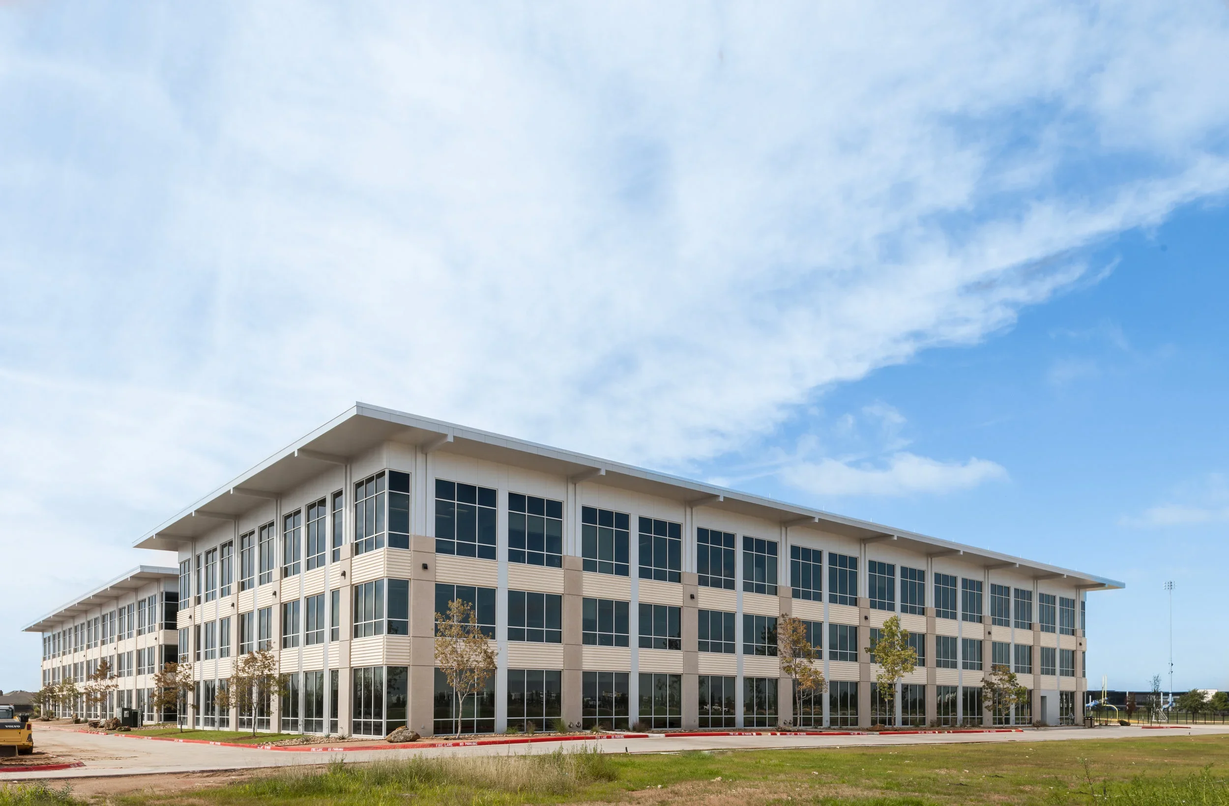 Modern multi-story office building with large windows, trees in front, and a parking lot under a partly cloudy sky.