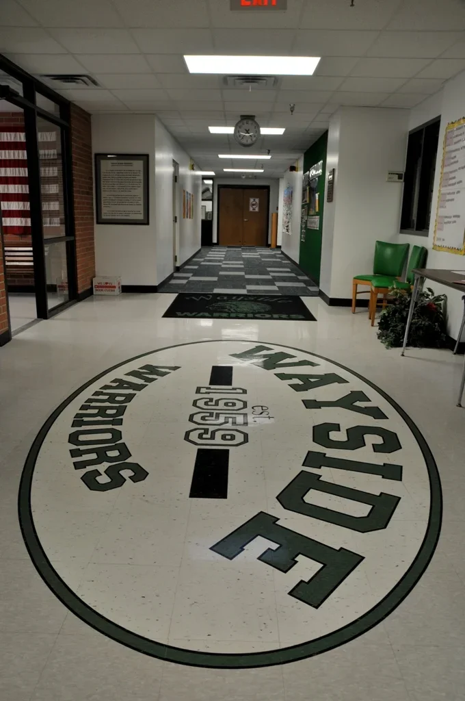 Indoor hallway with floor decal of the FAYETTEVILLE PUBLIC SCHOOL mascot, featuring a shield and the text "Fayetteville Public Schools".
