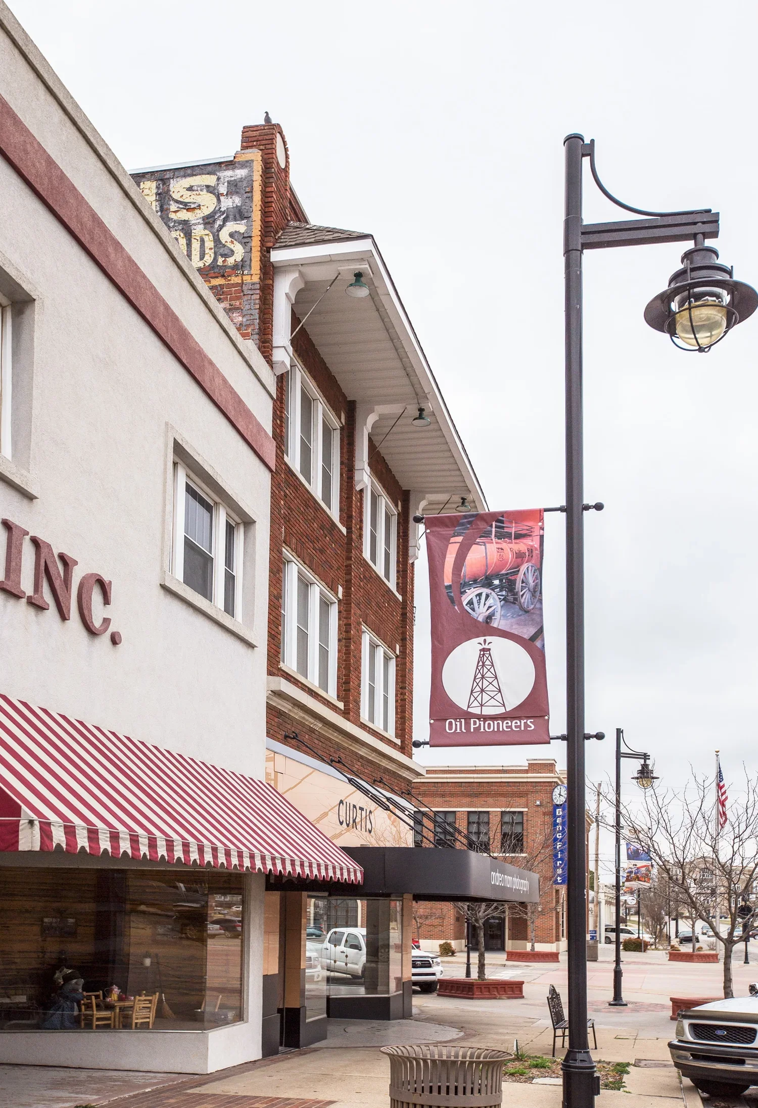 Street view of a downtown area with a building featuring a banner that reads 'Oil Pioneers' and an illustration of an oil derrick, with surrounding shops, a lamppost, parked cars, and leafless trees.
