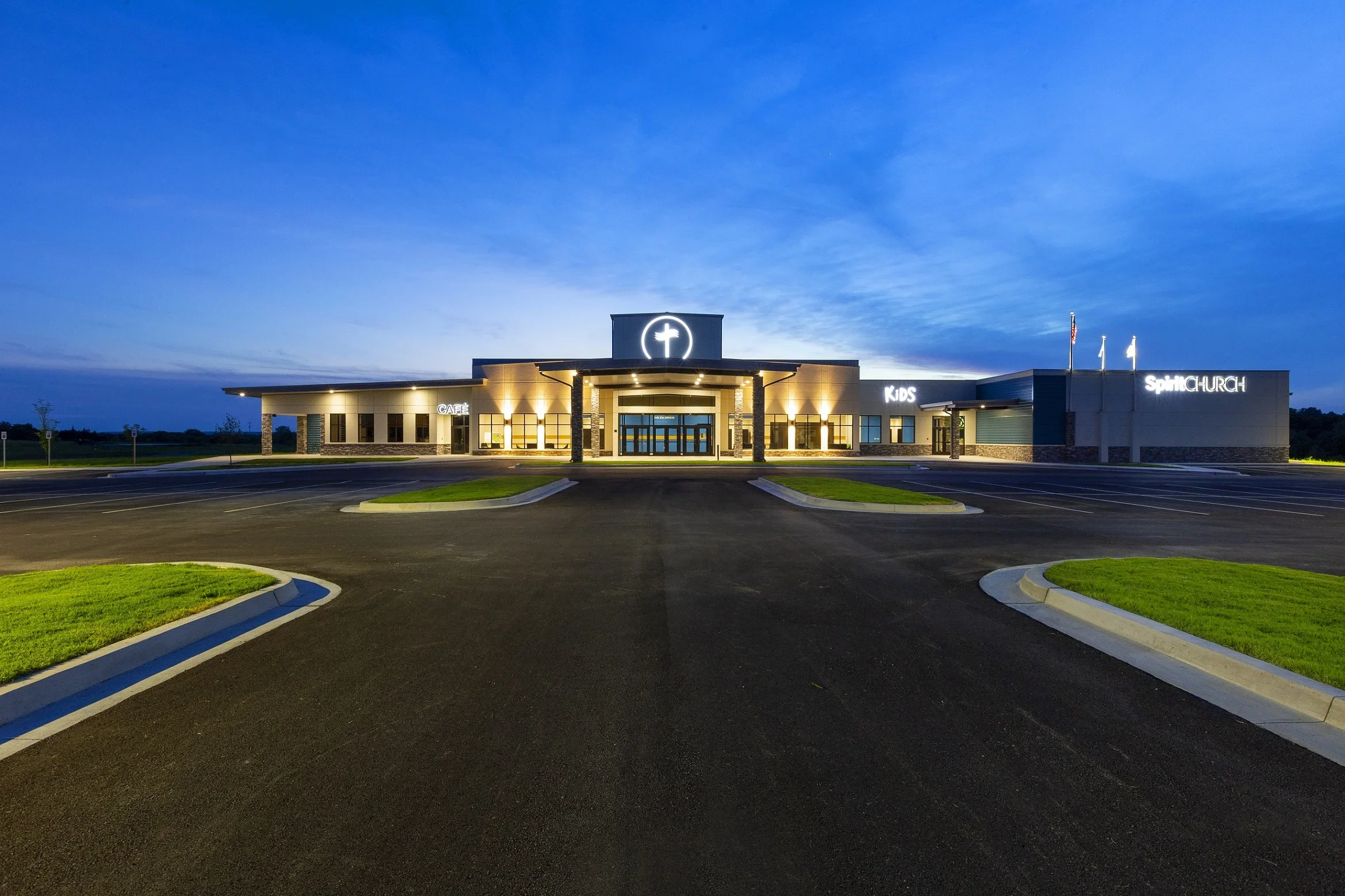 A modern church building illuminated at dusk with a large cross symbol on top, surrounded by an empty parking lot, with signs indicating areas for kids, café, and the main church entrance.
