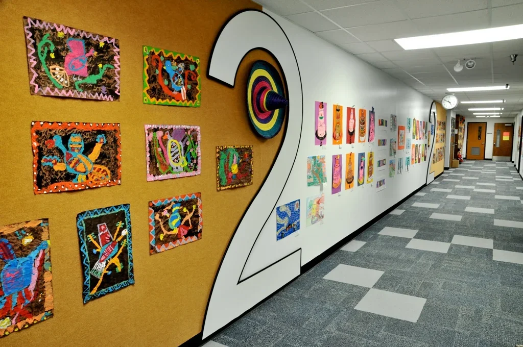 Colorful children's artwork displayed on a school hallway wall, with a large number 2 cutout in the center, and a clock on the wall in the background.