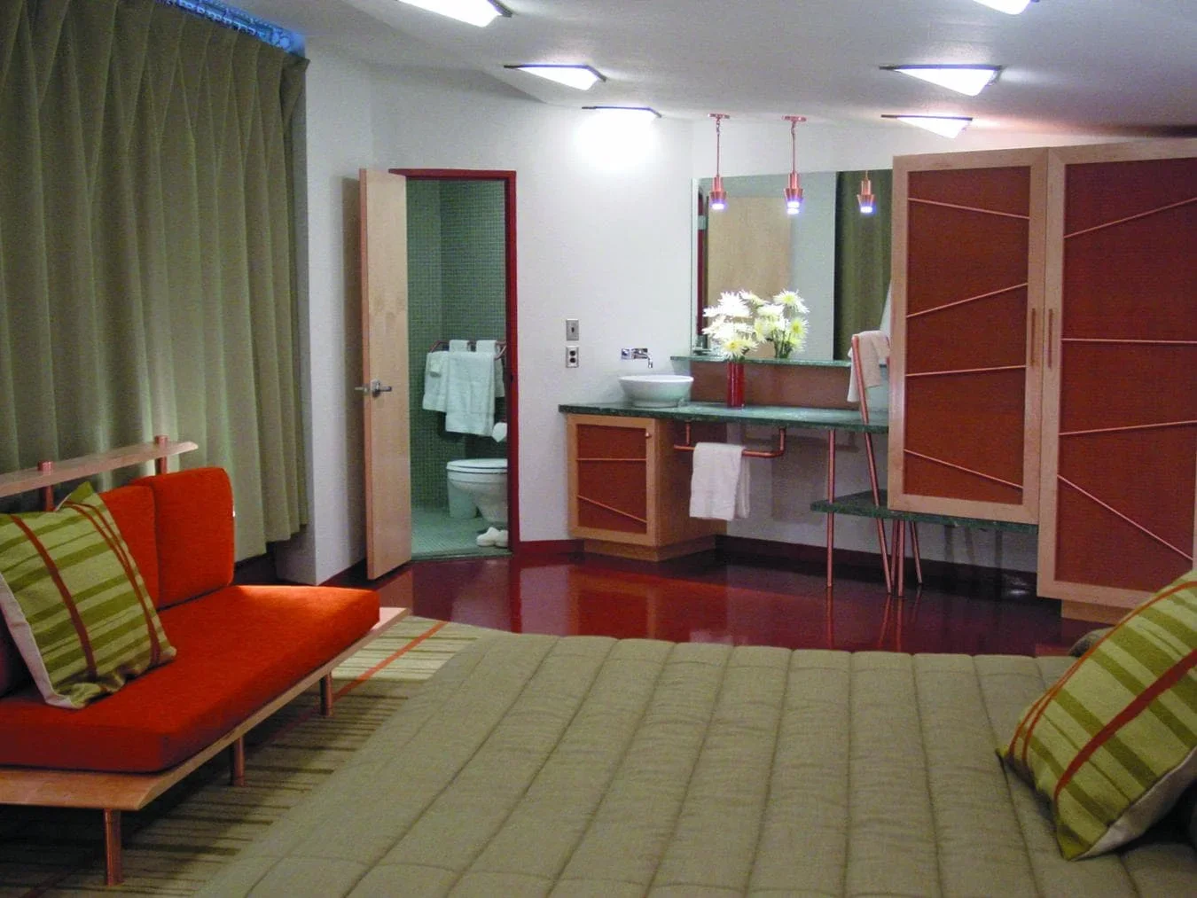 Hotel room with bed, orange and striped green pillows, open bathroom door, vanity with sink, mirror, and flowers, wooden cabinets, and curtains.