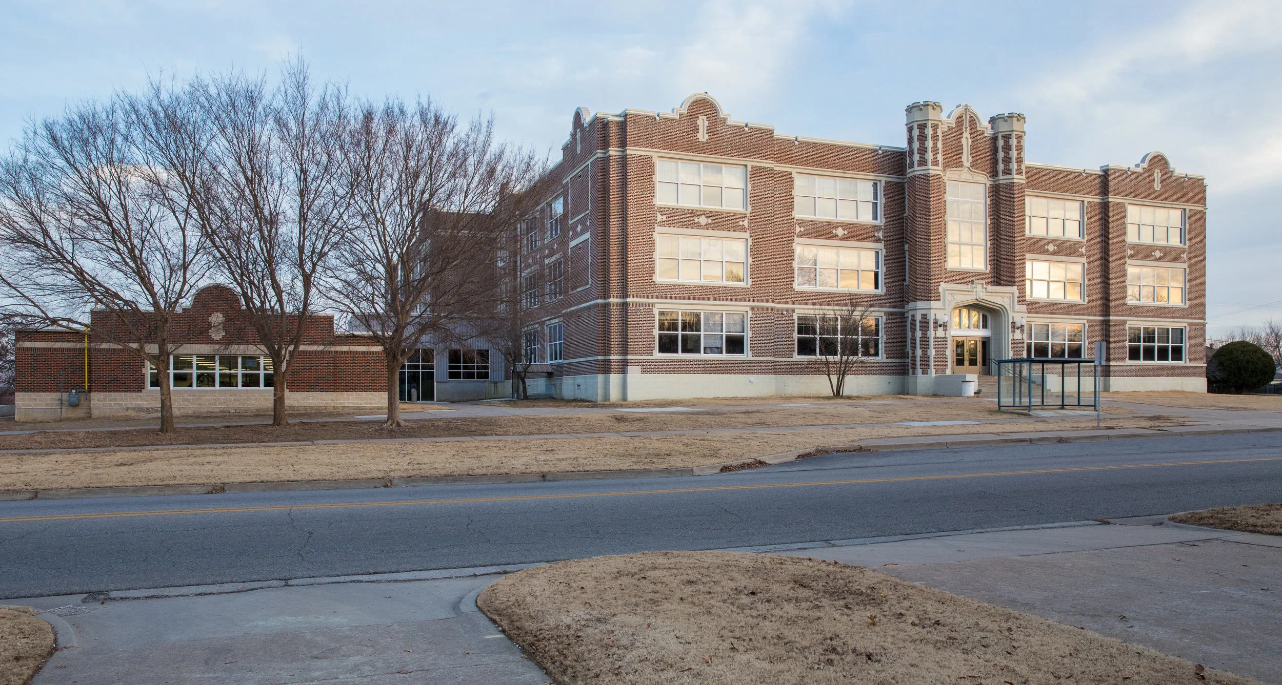 A large brick school building with four floors, decorative architectural details, and multiple windows. In front are leafless trees, a sidewalk, and a street.