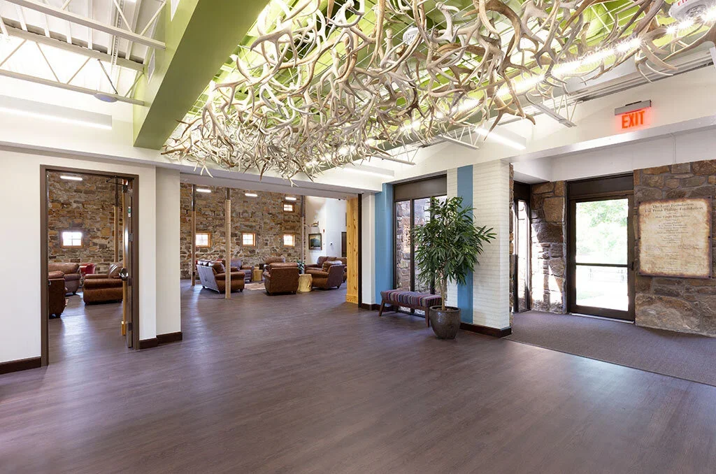 Lobby with a large hanging plant sculpture on the ceiling, potted plant near glass door, and seating area with brown chairs and stone walls.