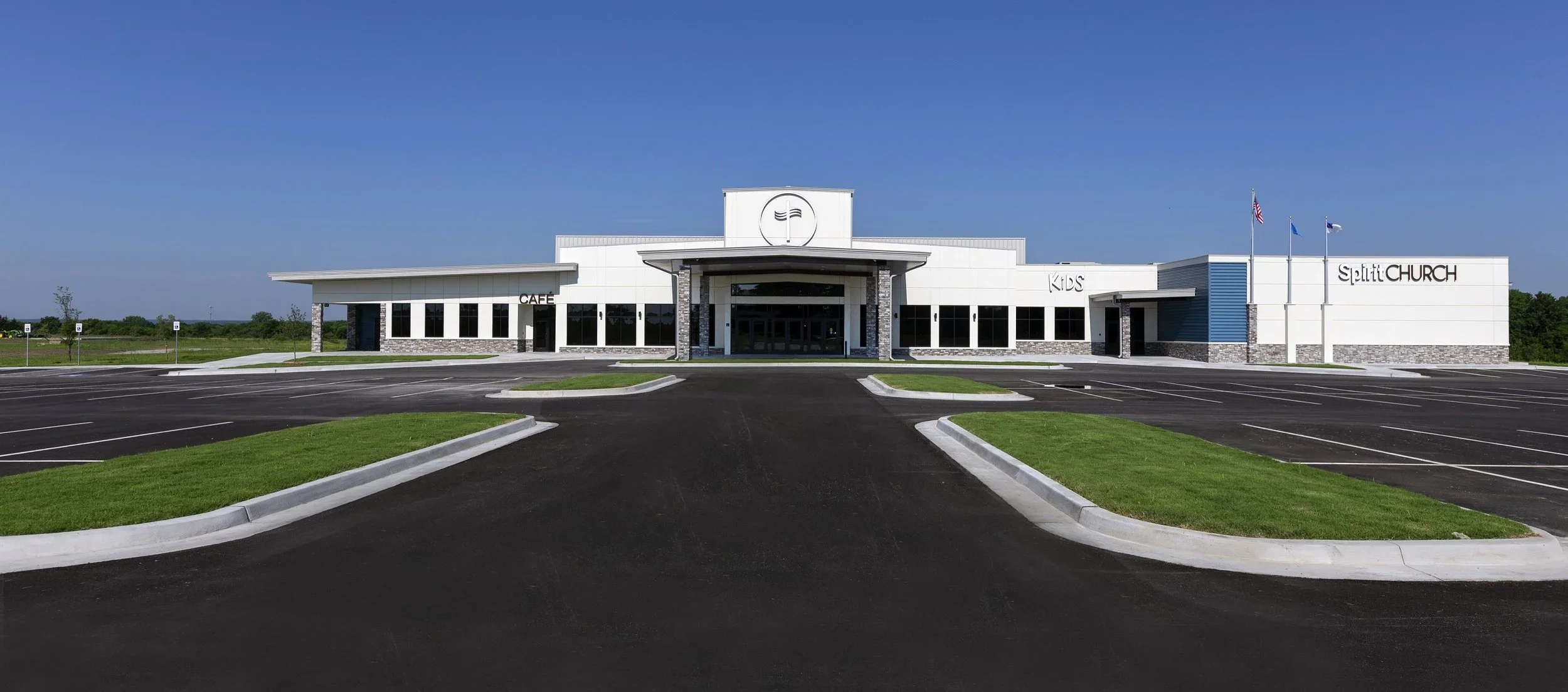Exterior view of a modern church building with parking lot, green grass patches, and clear blue sky.