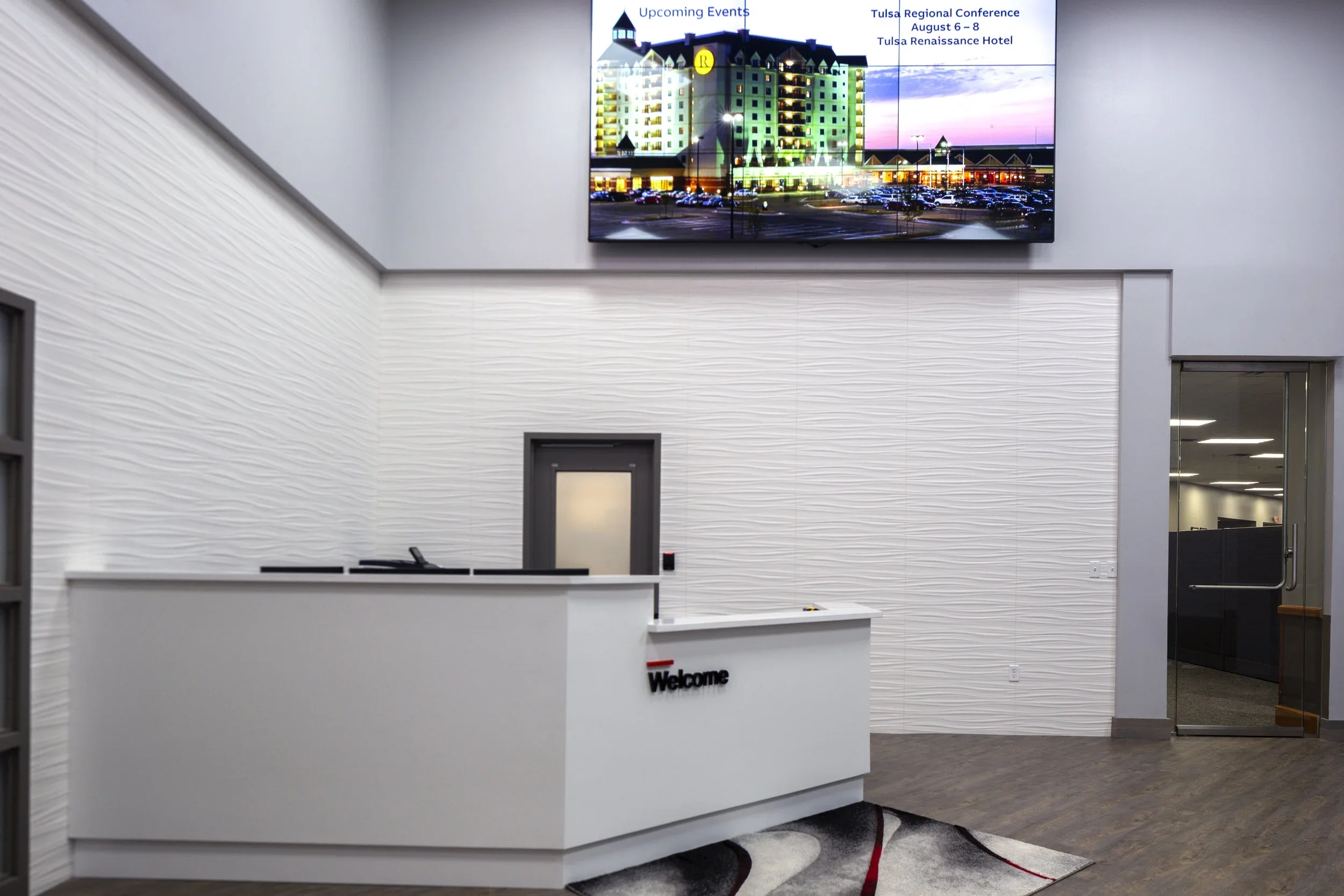 Hotel reception desk with a welcome sign, a door behind the desk, a large wall-mounted digital display showing a building and upcoming events, and an open glass door leading to an office area.